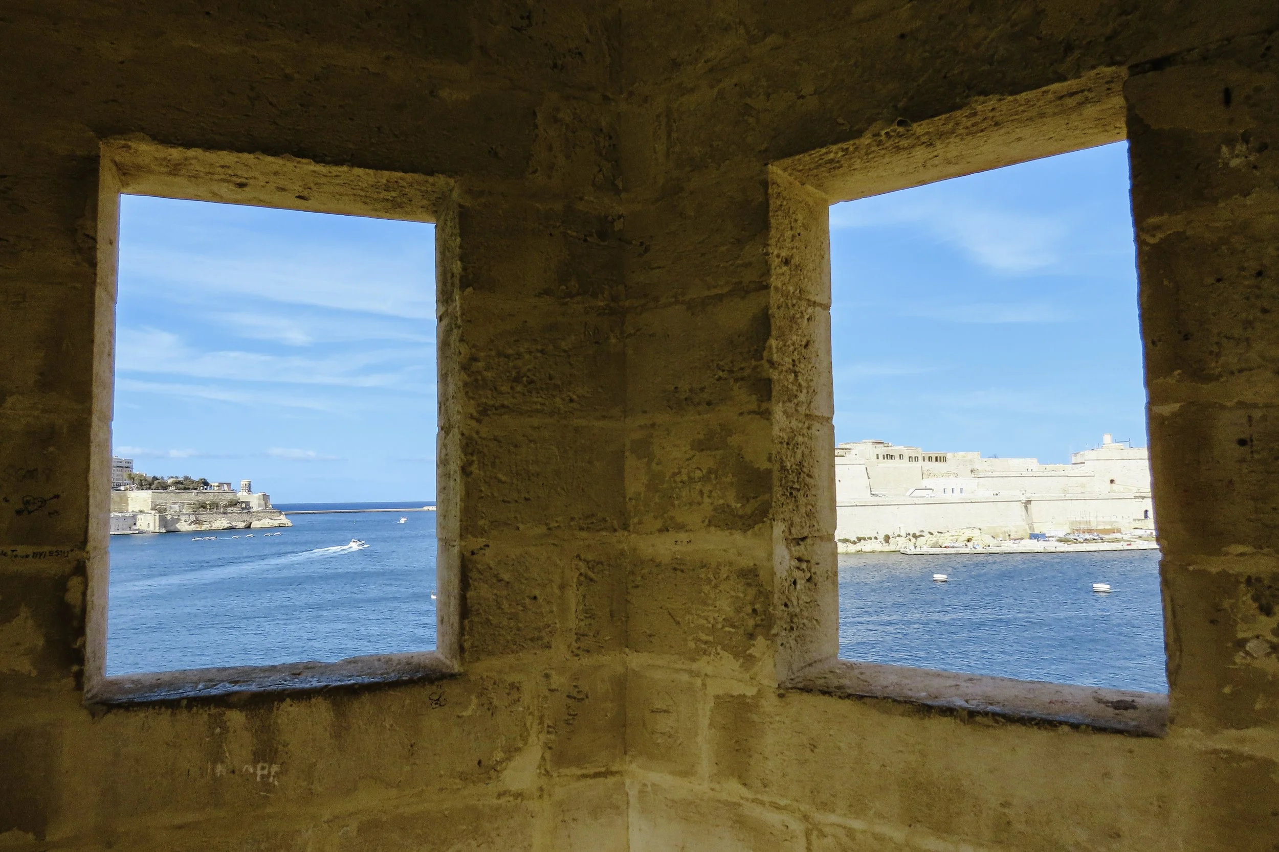 Vista del mare e di una fortezza attraverso due finestre in un muro di pietra.