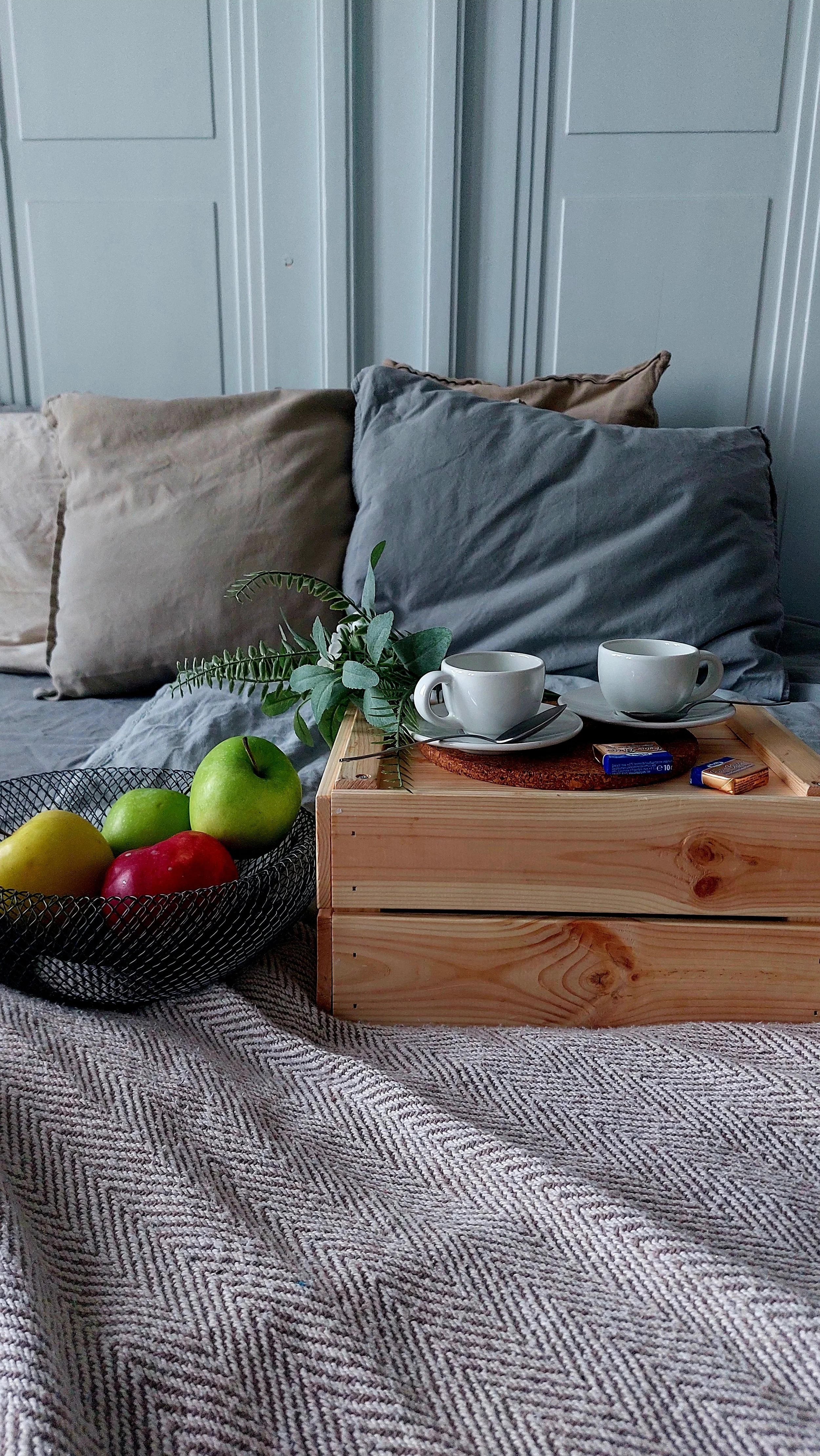 Un letto con cuscini beige e grigi, un vassoio di legno con due tazze e piattini, una pianta verde e una cesta con mele verdi e rosse. La scena è in stile minimalista con luci morbide.