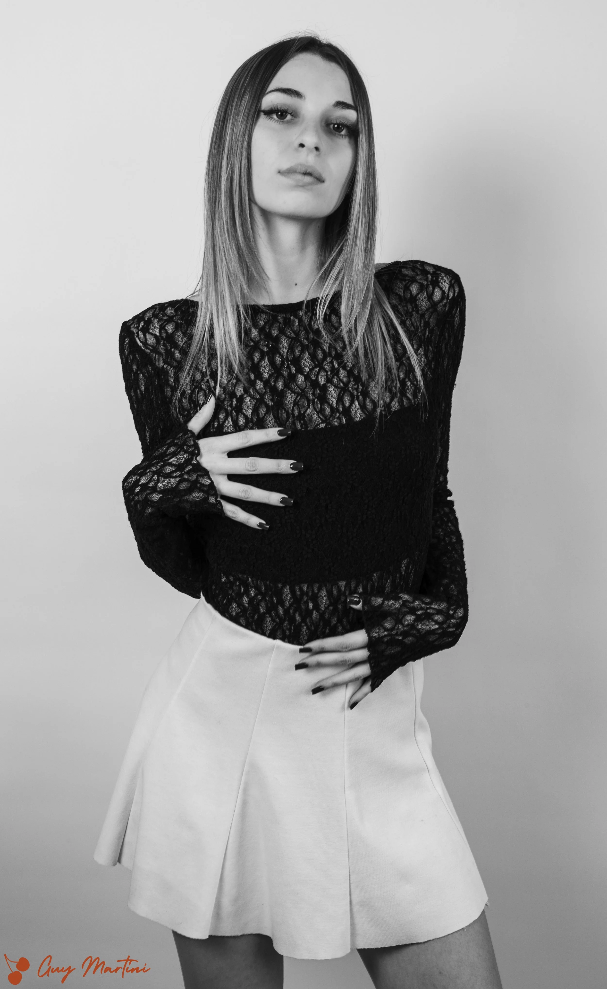 Ragazza con abito nero a maniche lunghe in pizzo, gonna beige, posa con le mani sul ventre e sul petto, capelli lunghi e lisci, sfondo bianco.