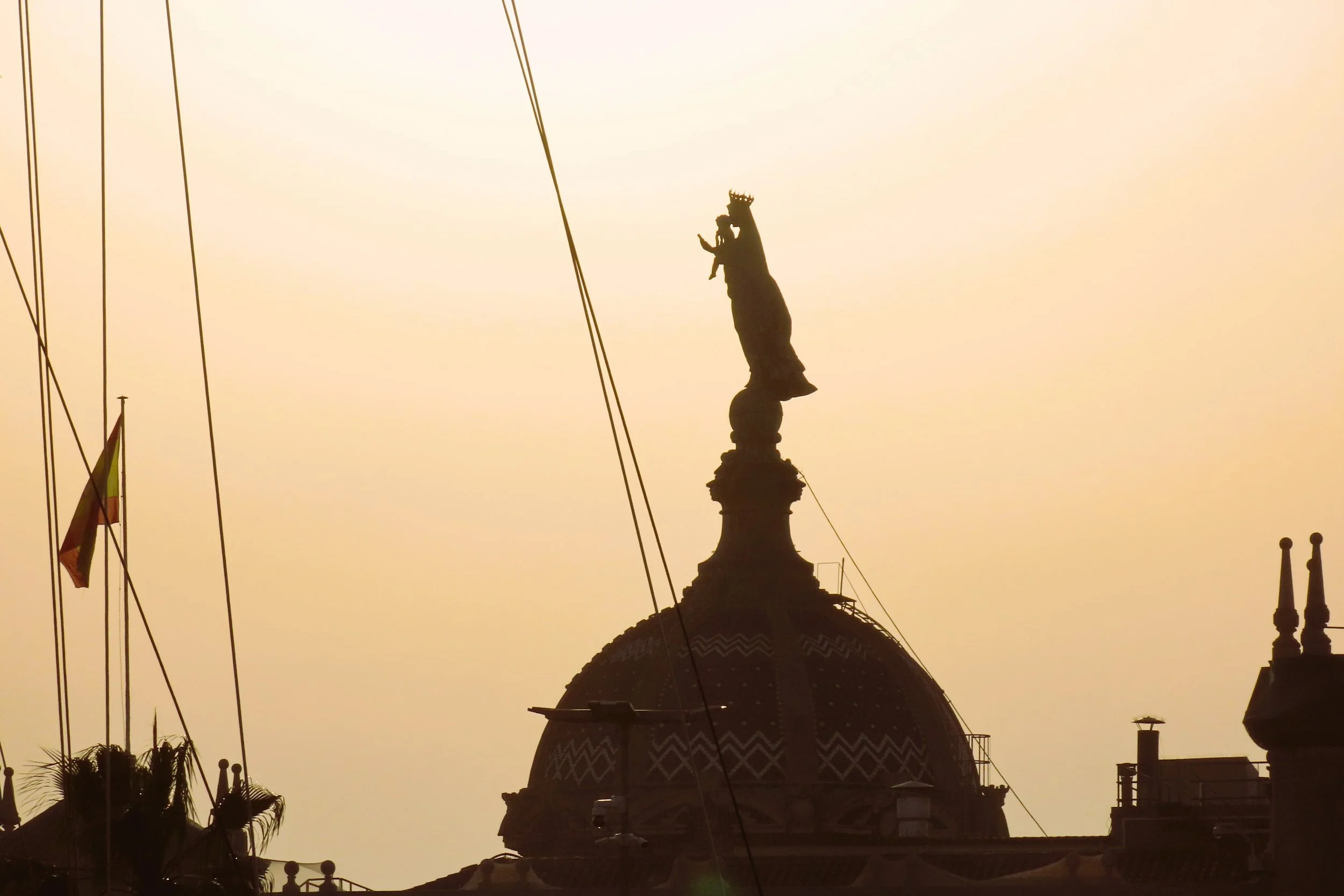 Silhouette di un edificio storico con una statua al centro di una cupola, bandiere al vento e un cielo al tramonto.