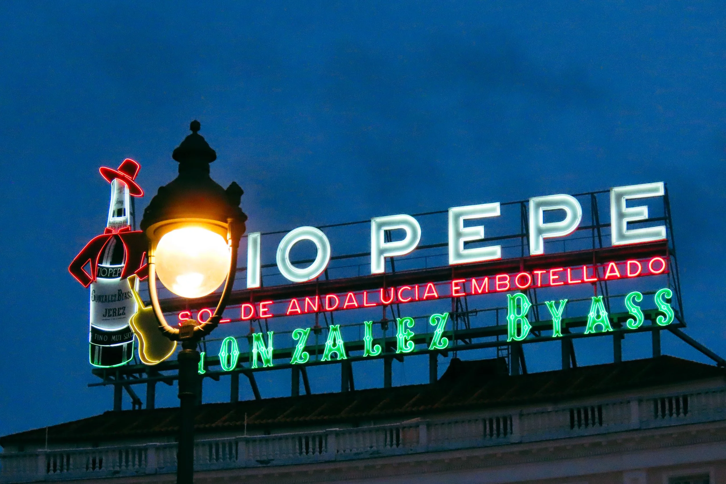 Segnale luminoso con il testo 'IO PEPE' e 'SOTTO DE ANDALUCIA EMBOTELLADO' e 'GONZALEZ BASS' sotto una lampada rossa, con un'illustrazione di una bottiglia di gin Gonzalez Bass e un cappello rosso.