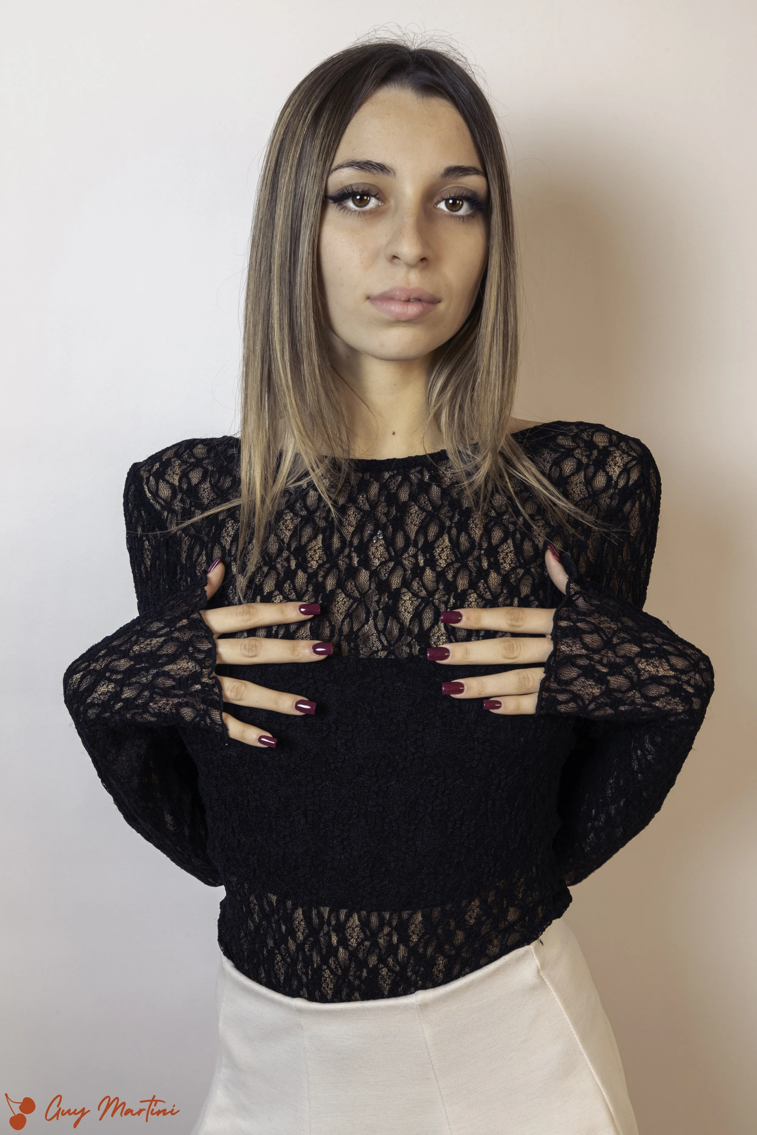 Ragazza con capelli lunghi, vestita con un maglione nero in pizzo e una gonna beige, che guarda in camera con espressione seria.