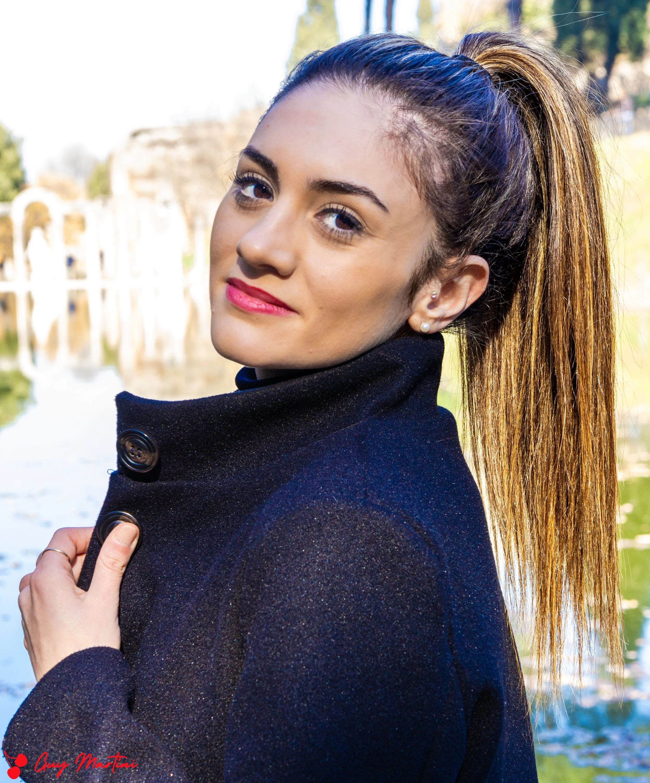 Ragazza con capelli castani lunghi in una coda alta, indossa un cappotto nero e sorride davanti a uno sfondo di acqua e natura.