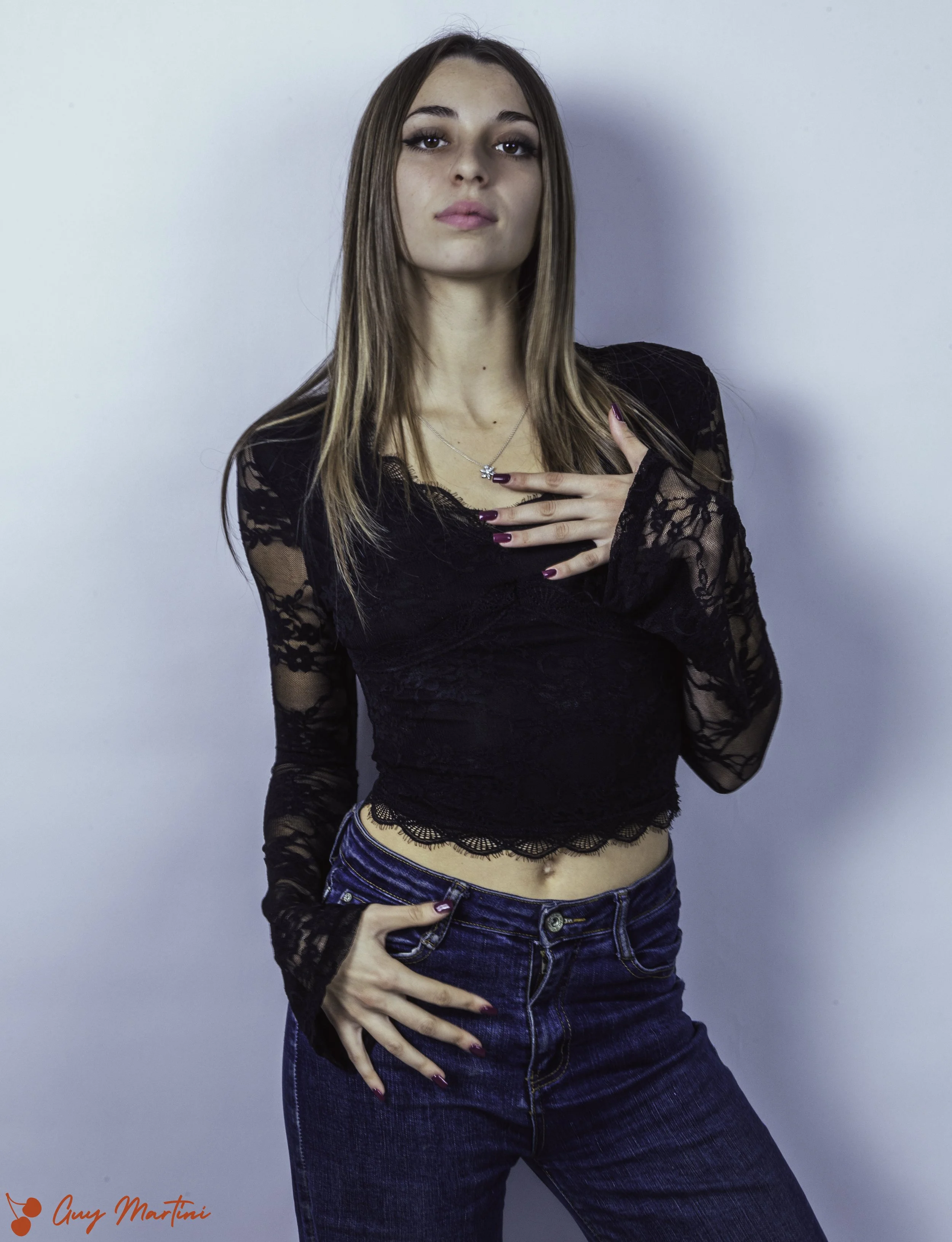 Una giovane donna con capelli lunghi castani, indossa una maglietta di pizzo nero e jeans blu, posa contro uno sfondo chiaro.