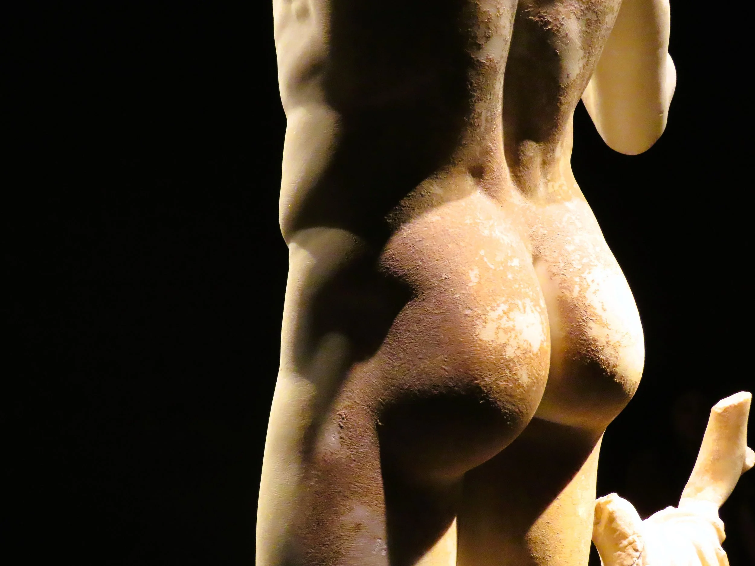 Particolare di una statua in marmo che rappresenta un sedere femminile con un braccio e una mano visibili, illuminata da una luce calda contro uno sfondo nero.