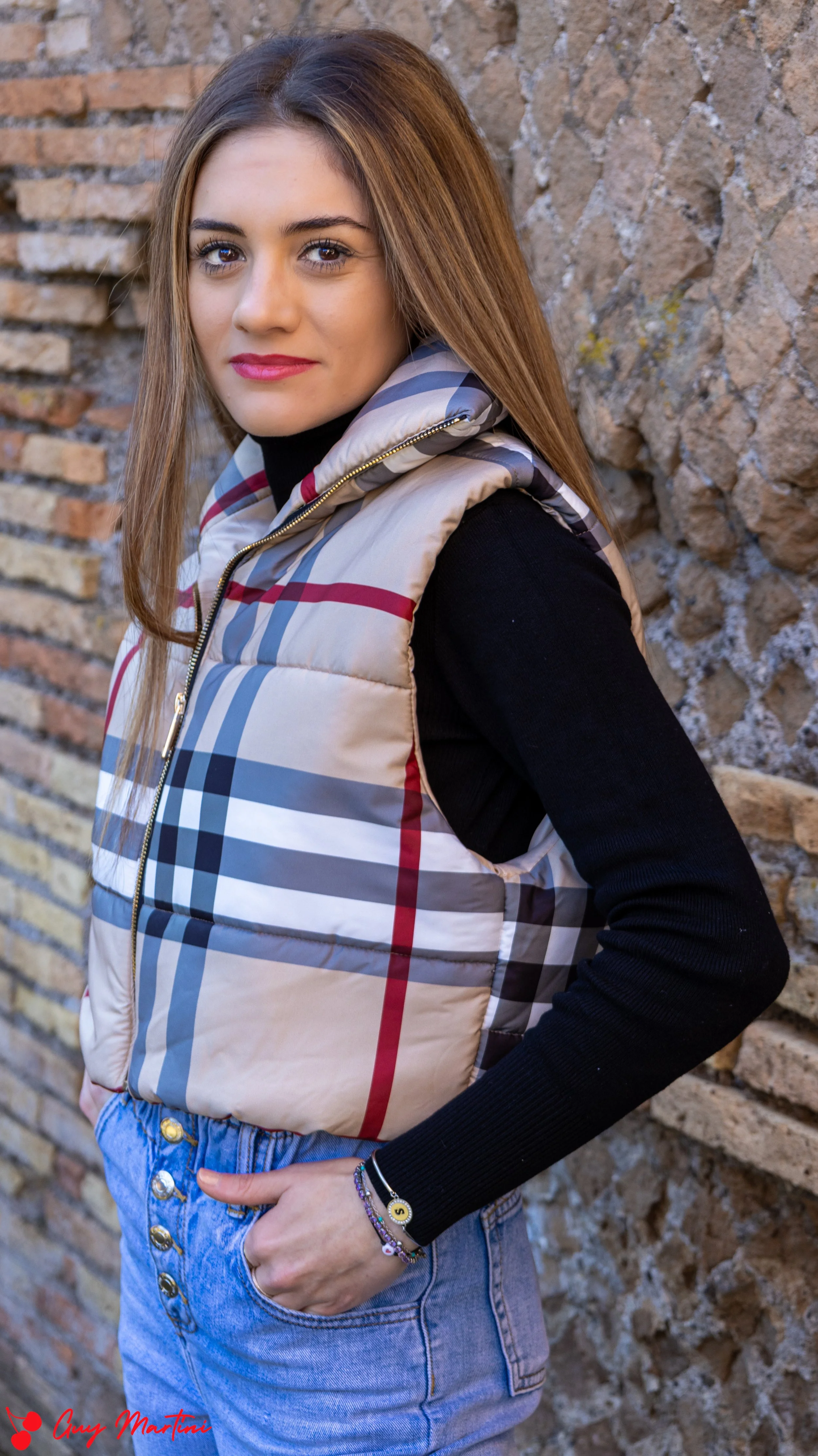 Ragazza con giubbotto imbottito con motivo a scacchi beige, nero, rosso e blu, pantaloni jeans a vita alta, posa con una mano in tasca, sfondo di un muro di pietre.