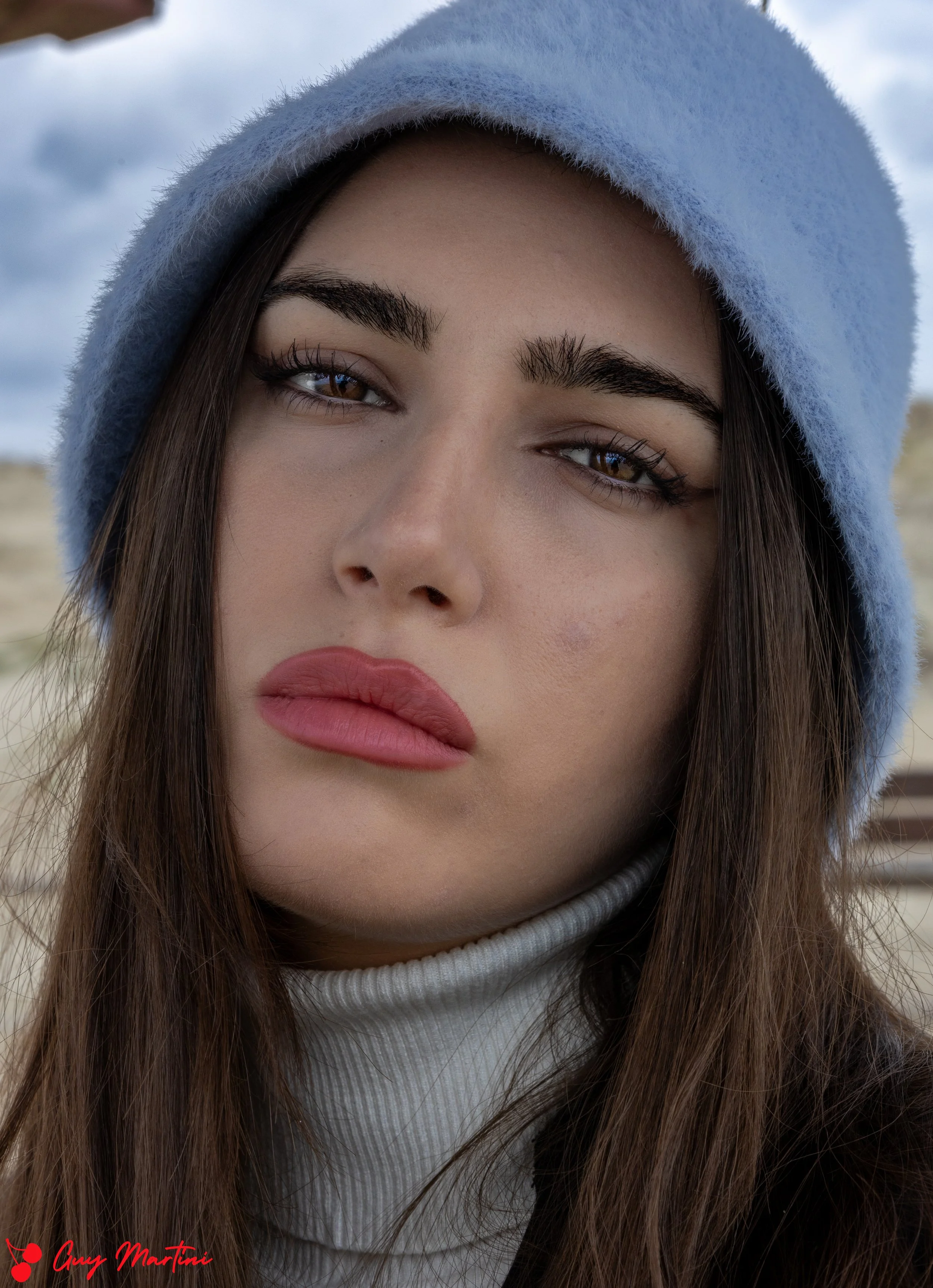 Ragazza con cappello blu all'aperto, volto in primo piano, espressione seria, capelli castani lunghi, trucco naturale, sfondo nuvoloso.