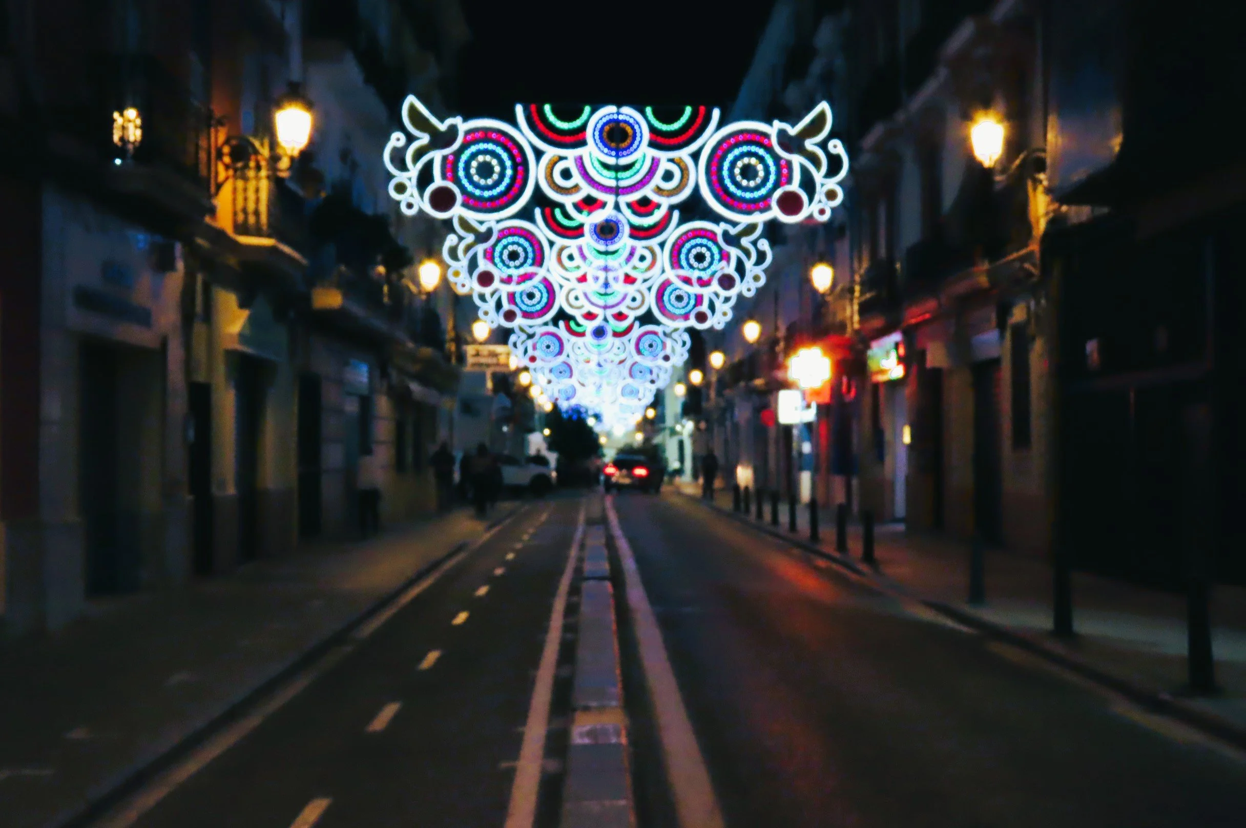 Strada di sera decorata con luci natalizie colorate a forma di disegni astratti e cerchi, illuminando la via con edifici e lampioni accesi.