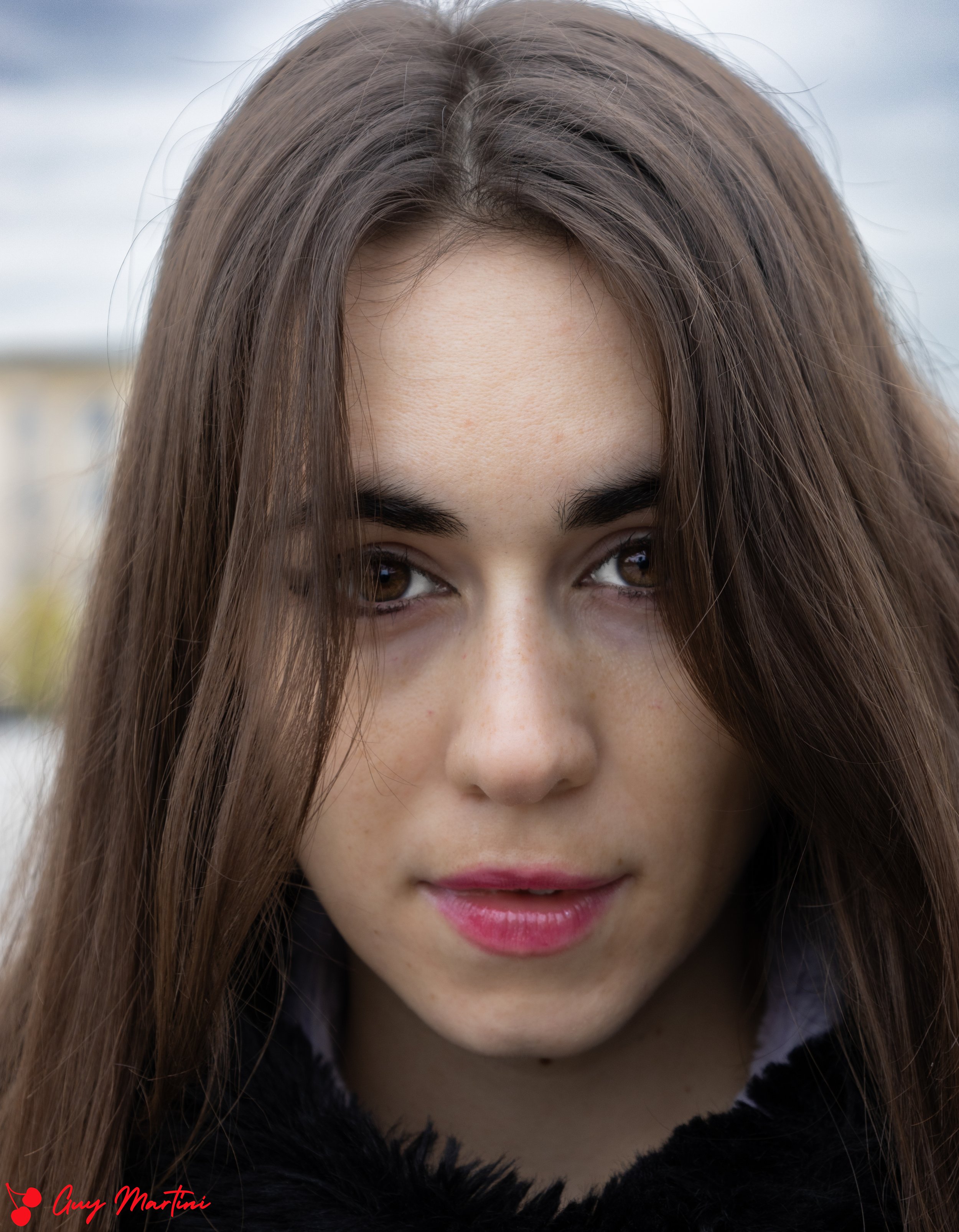  Primo piano di una ragazza con capelli castani, occhi castani, labbra rosa e pelle chiara con lentiggini, con sfondo di cielo nuvoloso. 