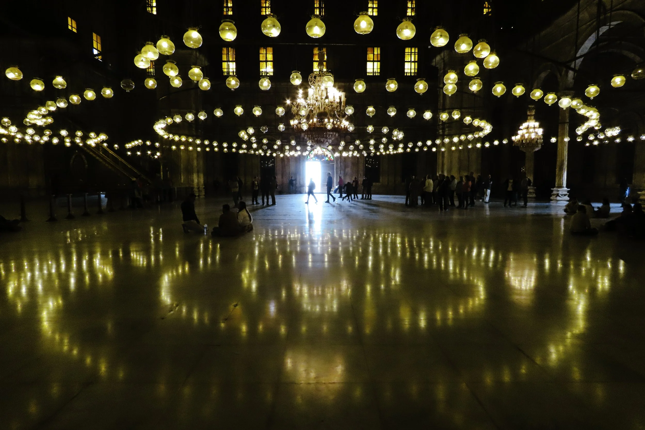 Interno di una grande sala con luci a chandelier, persone sedute e in piedi, e un'apertura con luce naturale che crea un riflesso sul pavimento.