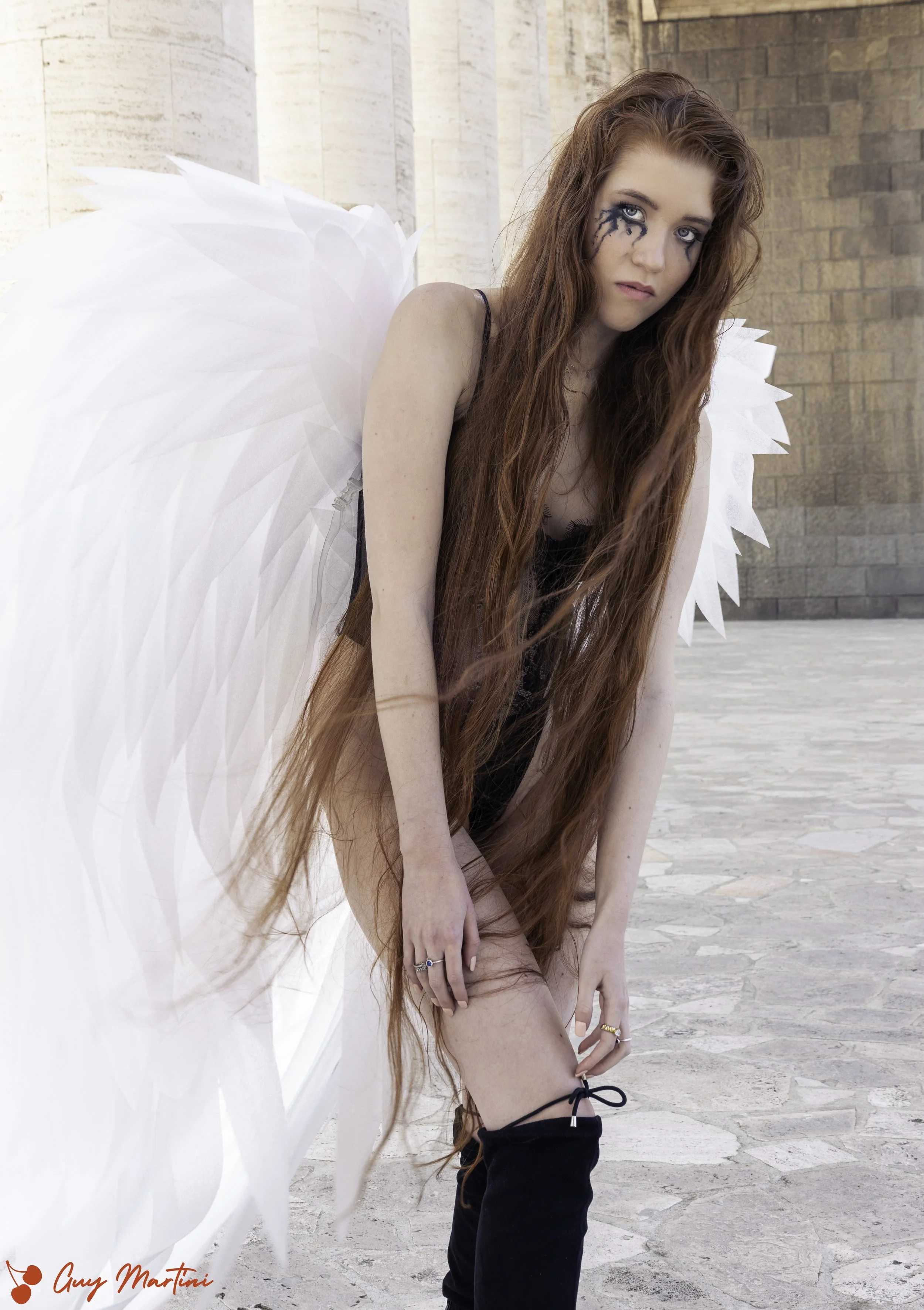 Donna con lunghi capelli rossi e ali da angelo bianche, vestita con abbigliamento nero, che si mette una calza nera mentre si piega in avanti.