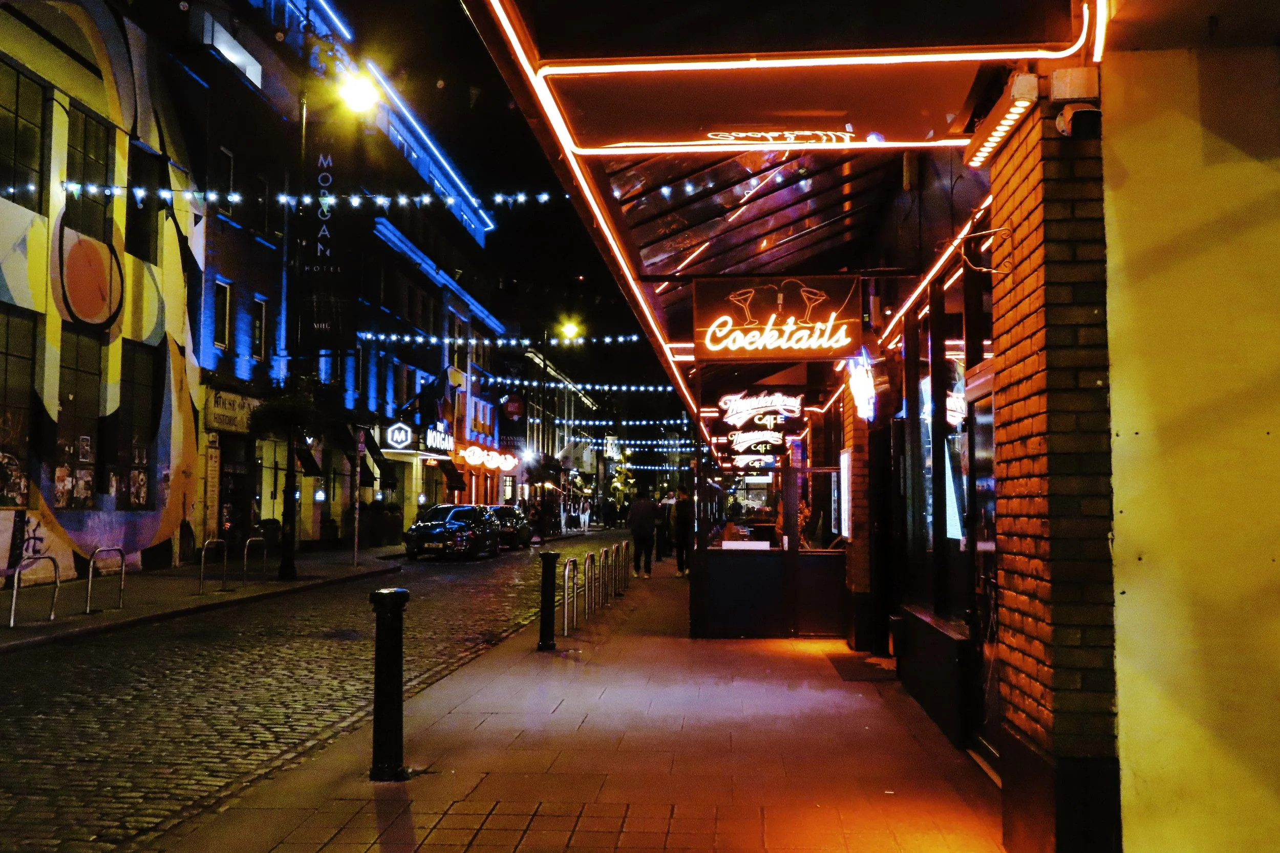 Strada cittadina con negozi e locali, luci al neon rosse e blu, con segnaletica per cocktail e caffè, e decorazioni luminose sulla strada durante la sera.