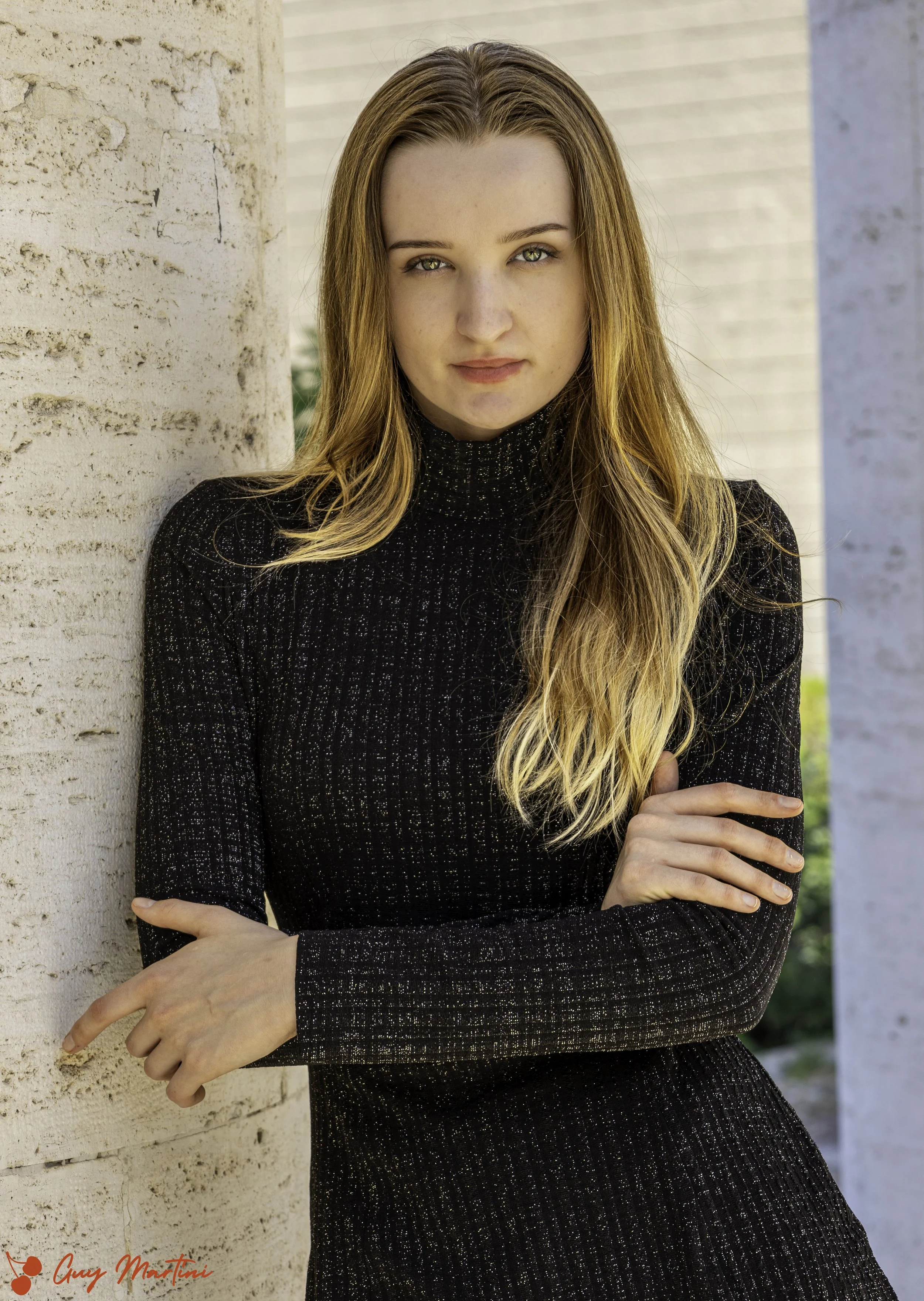 Ragazza con capelli castani e occhi chiari si appoggia a una colonna di pietra con un abito nero a maniche lunghe.