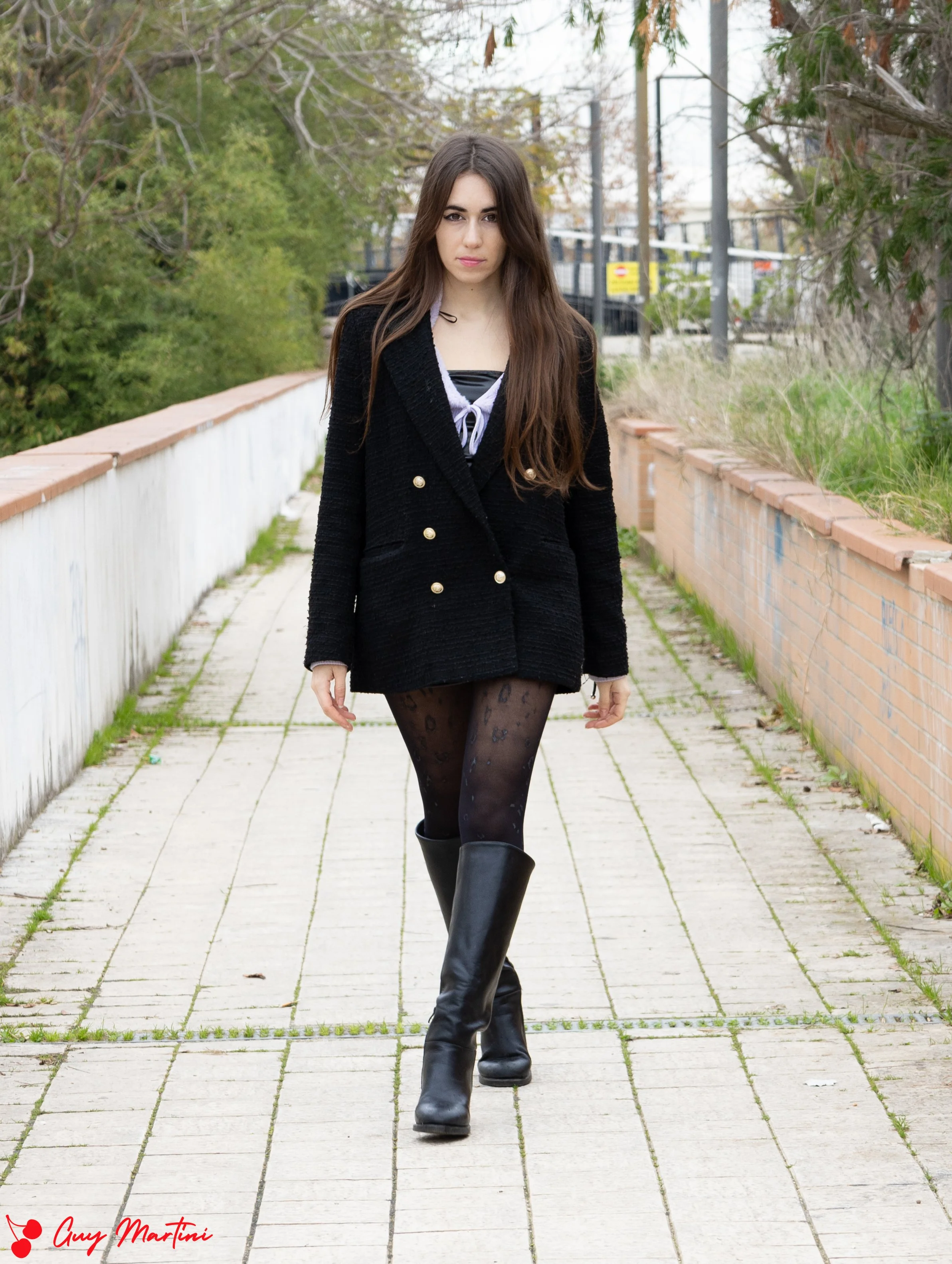 Una giovane donna con capelli lunghi e castani cammina lungo un percorso all'aperto, indossando un cappotto nero con bottoni dorati, calze nere con motivo leopardato e stivali alti neri.
