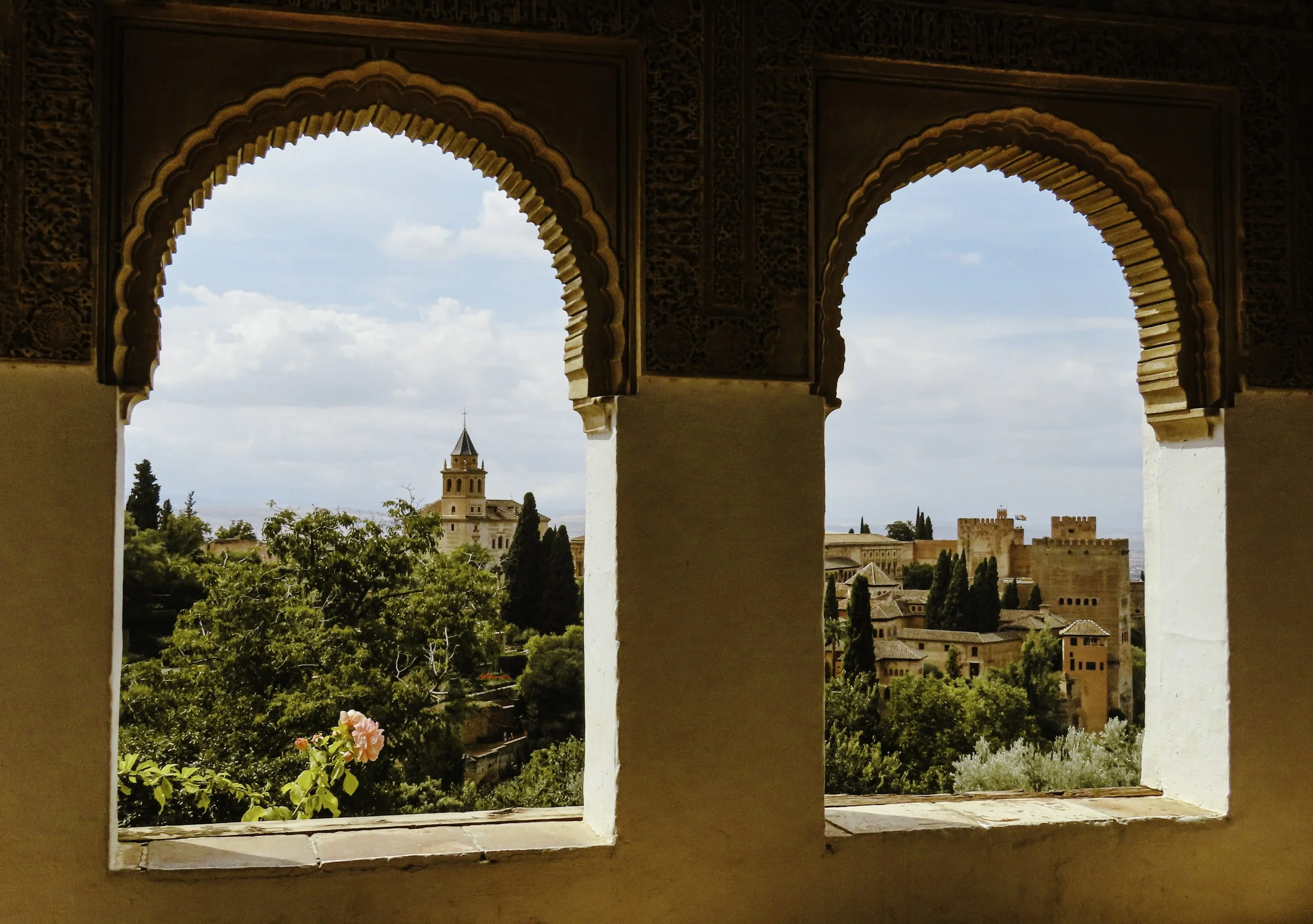 Vista del centro storico di Granada attraverso due archi decorati, con alberi e edifici storici, incluso un campanile, sotto un cielo nuvoloso.