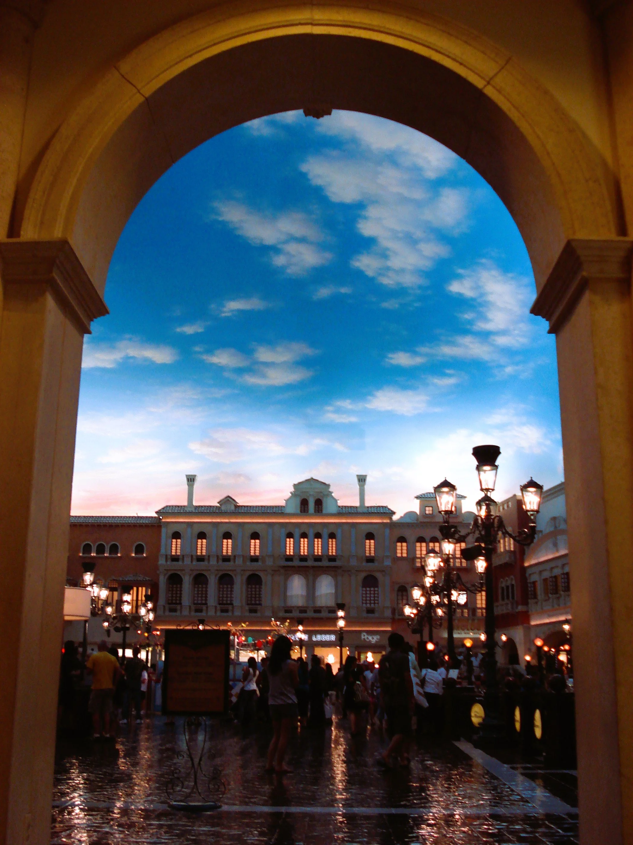 Veduta di un'architettura in stile europeo con edifici a più piani e illuminazione calda, sotto un cielo sereno e nuvoloso, con persone che passeggiano in una piazza durante la sera.