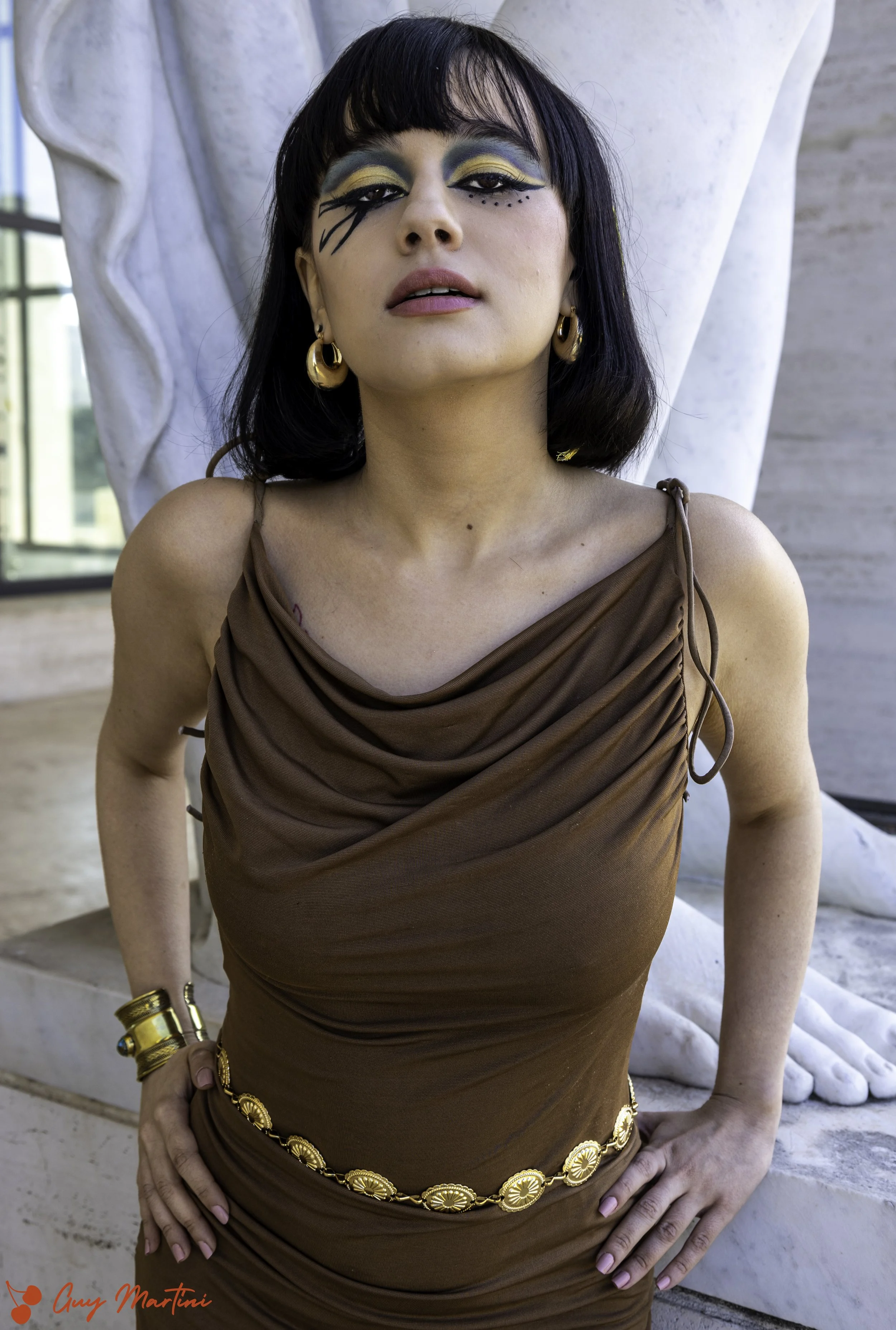 Donna con trucco artistico sugli occhi, indossa un vestito marrone con drappeggio e accessori dorati, posizione davanti a una scultura di marmo.