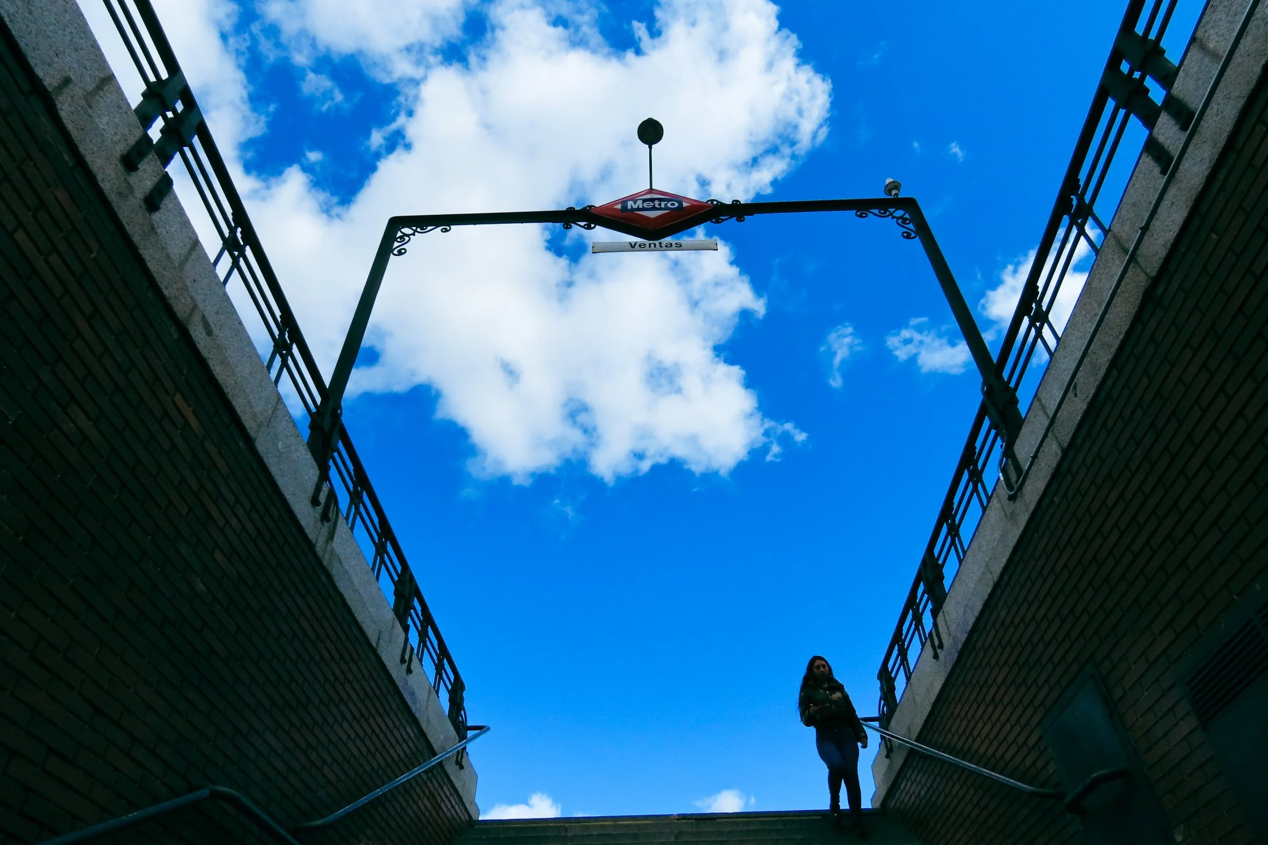 Una donna in piedi su una rampa di scale all'aperto in un'area urbana tra due pareti di mattoni, con il cielo blu e nuvole bianche sopra di lei.