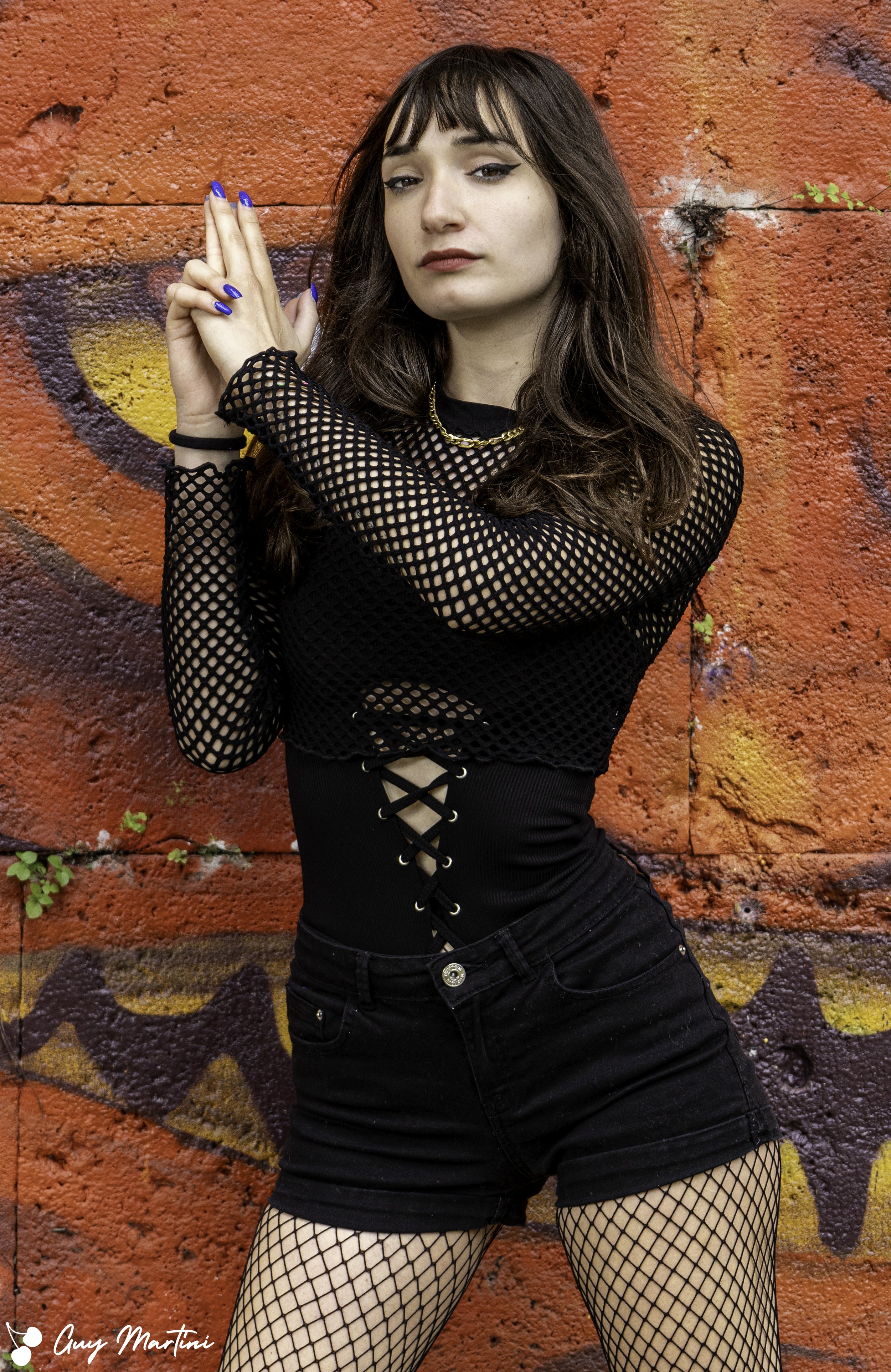 Ragazza con capelli lunghi e ricci, indossa un top nero con maniche in rete e shorts neri, posa contro un muro graffiti rosso e viola.