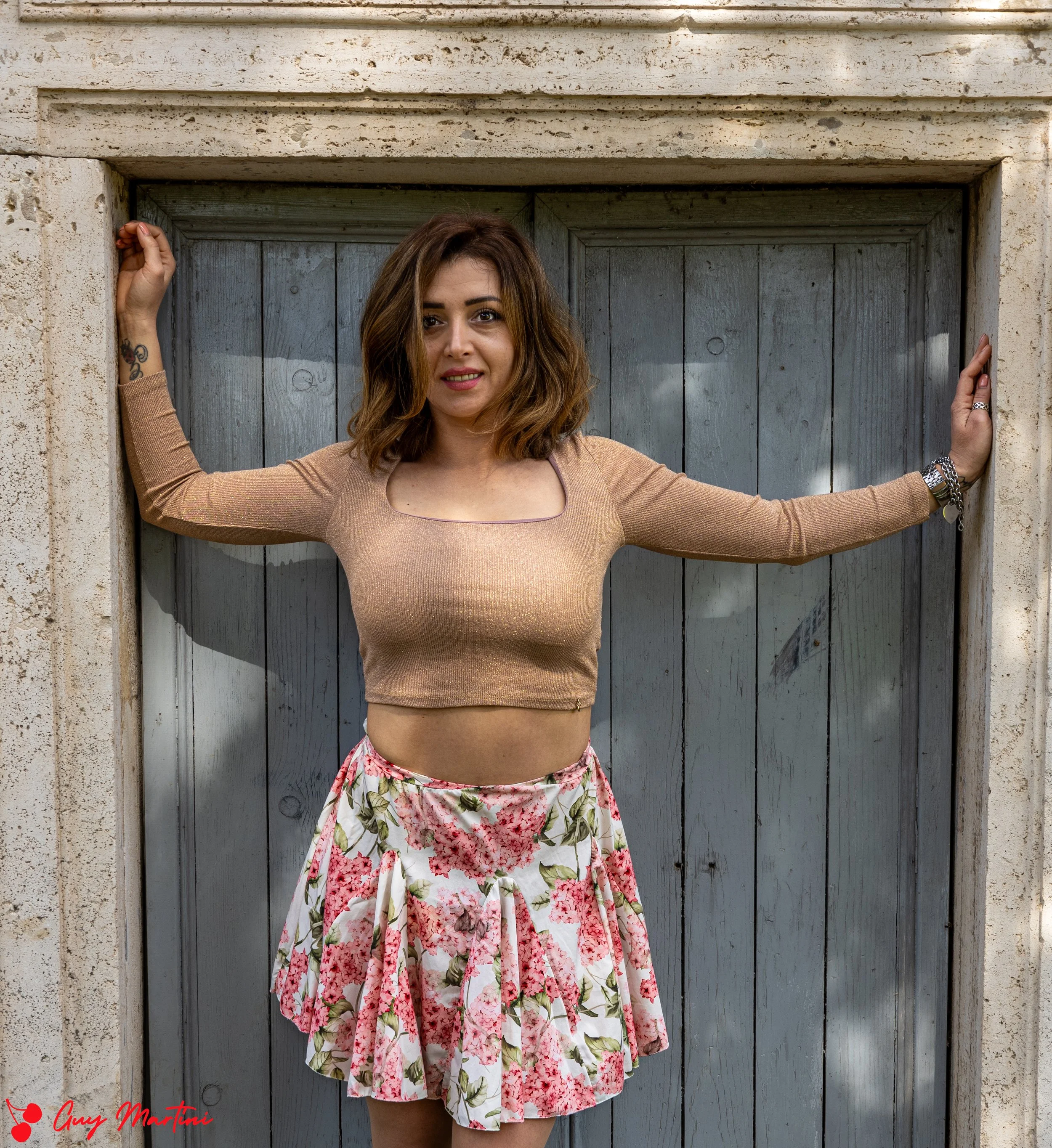 Una donna con capelli castani ricci, indossa una maglietta color oro e una gonna a fiori rosa e verde, si appoggia con le braccia alle pareti di una porta di legno grigia.