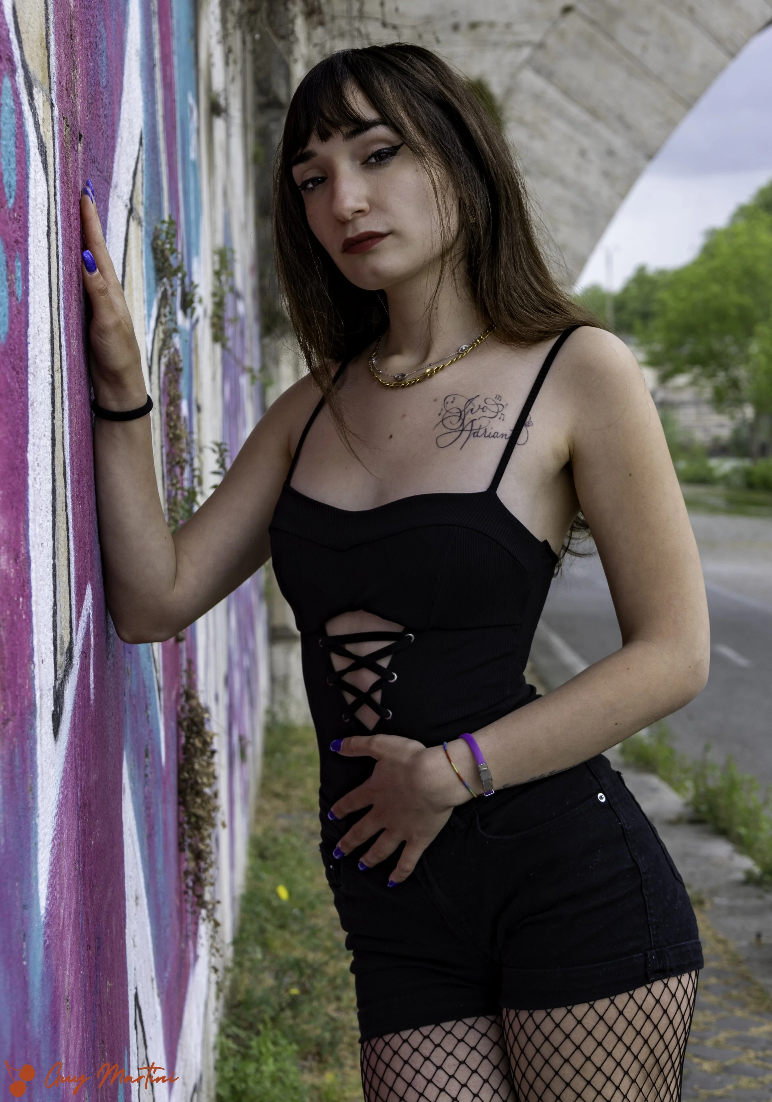 Una ragazza con capelli castani e vestito nero con bretelle e lacci al centro, posa contro un muro di graffiti colorati all'aperto, con un cielo nuvoloso e alberi sullo sfondo.