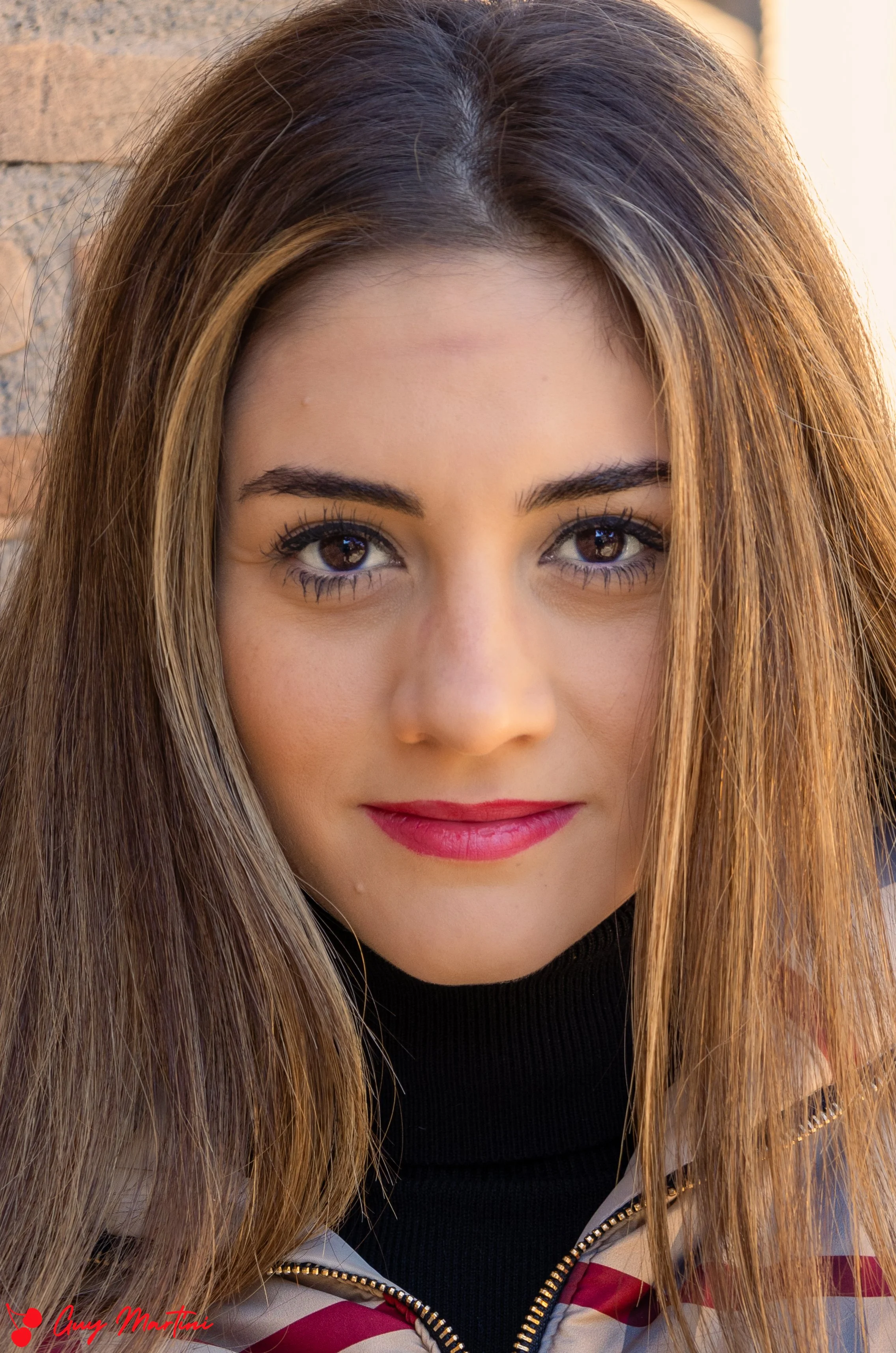 Ragazza con capelli castani, occhi grandi, sorriso con rossetto rosso, indossa una giacca con zip e sfondo di mattoni.