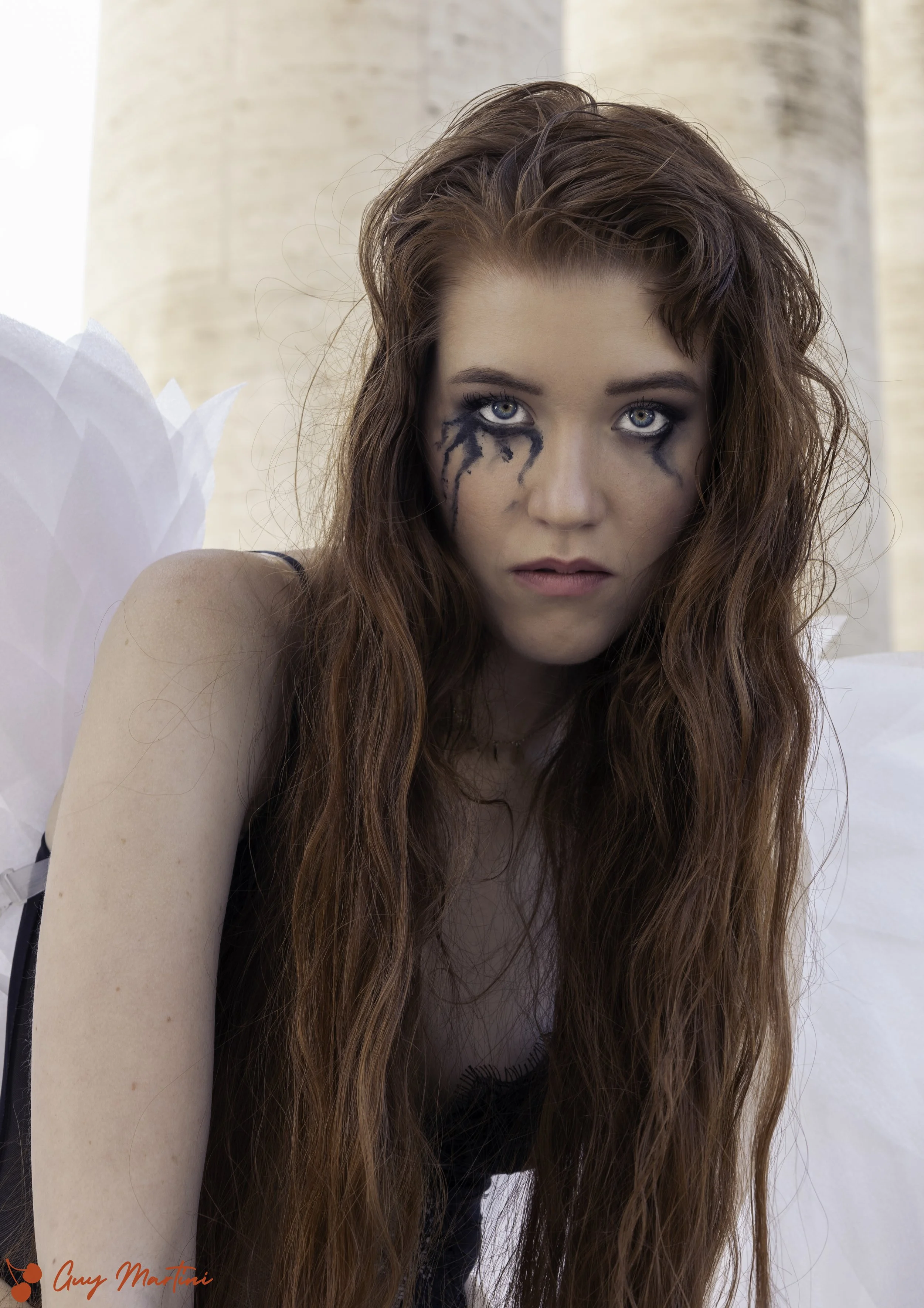 Ragazza con trucco occhi drammatico e capelli ricci rossi, con ali di carta sullo sfondo, posa in esterno.