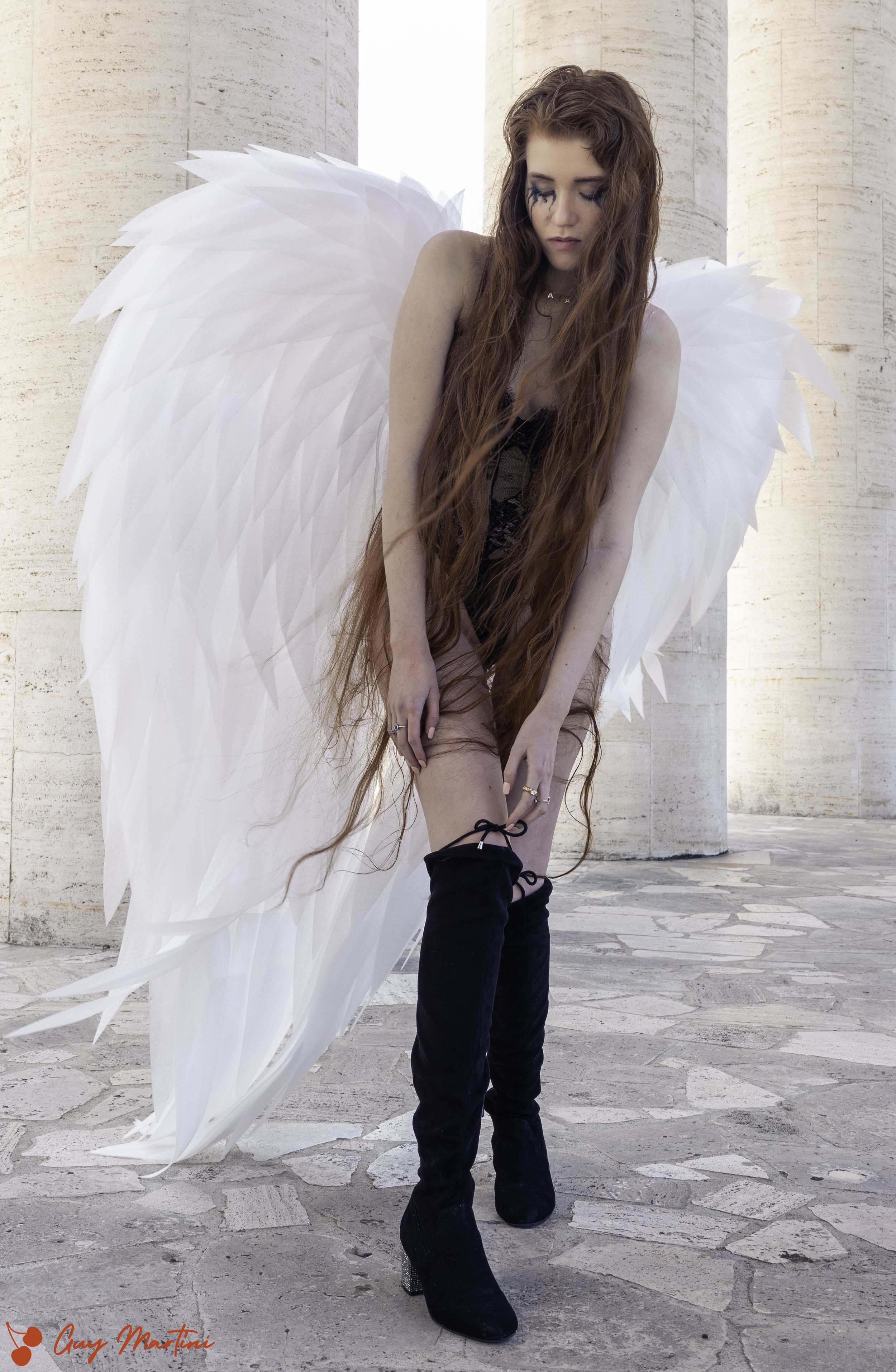 Una ragazza con lunghi capelli ricci scoloriti, abbigliamento nero, scarpe sopra il ginocchio, con grandi ali bianche e un ambiente esterno con pilastri di pietra.