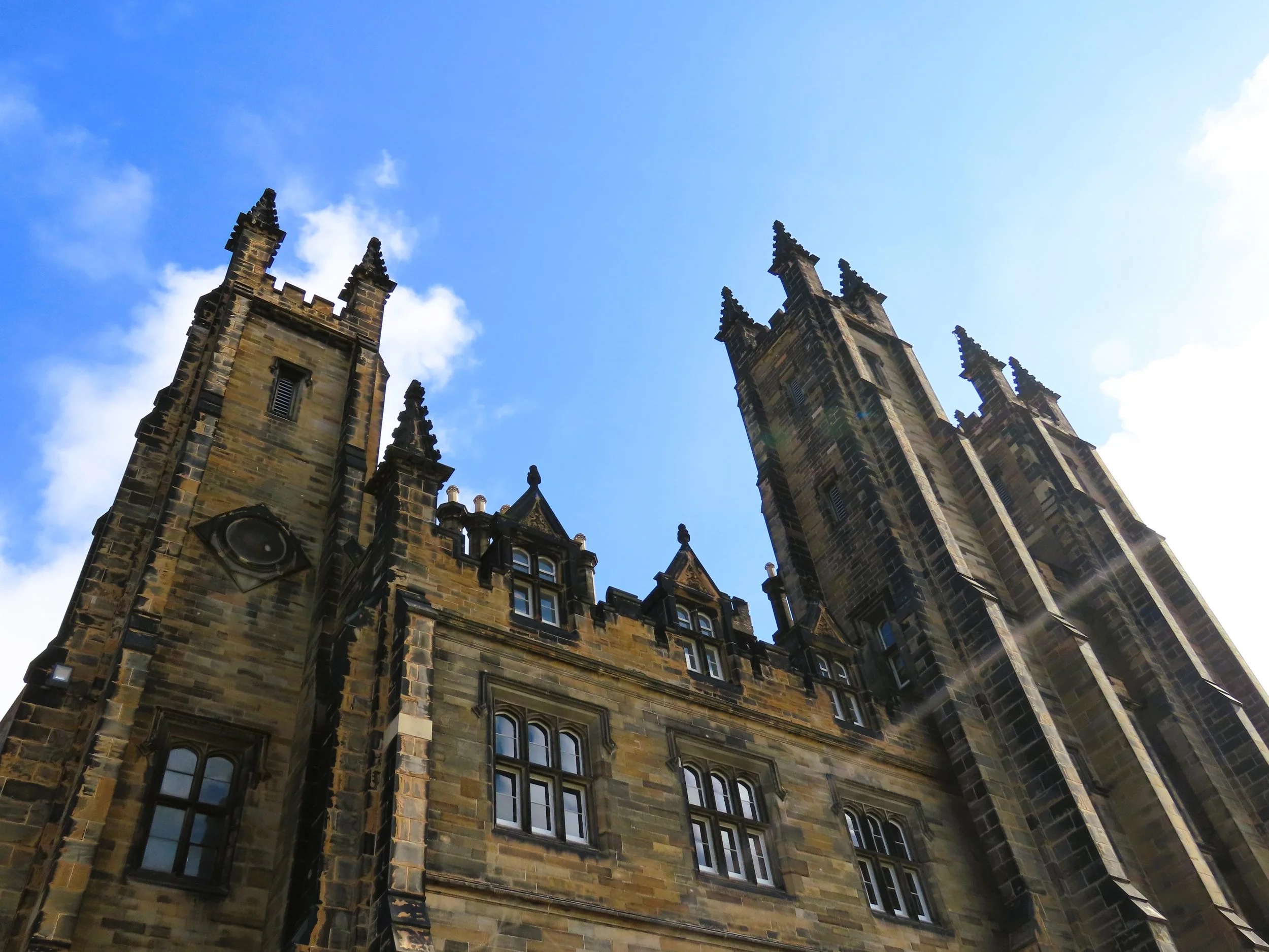 Veduta di due torri di un castello medievale in pietra, con dettagli architettonici gotici, sotto un cielo azzurro con nuvole sparse.