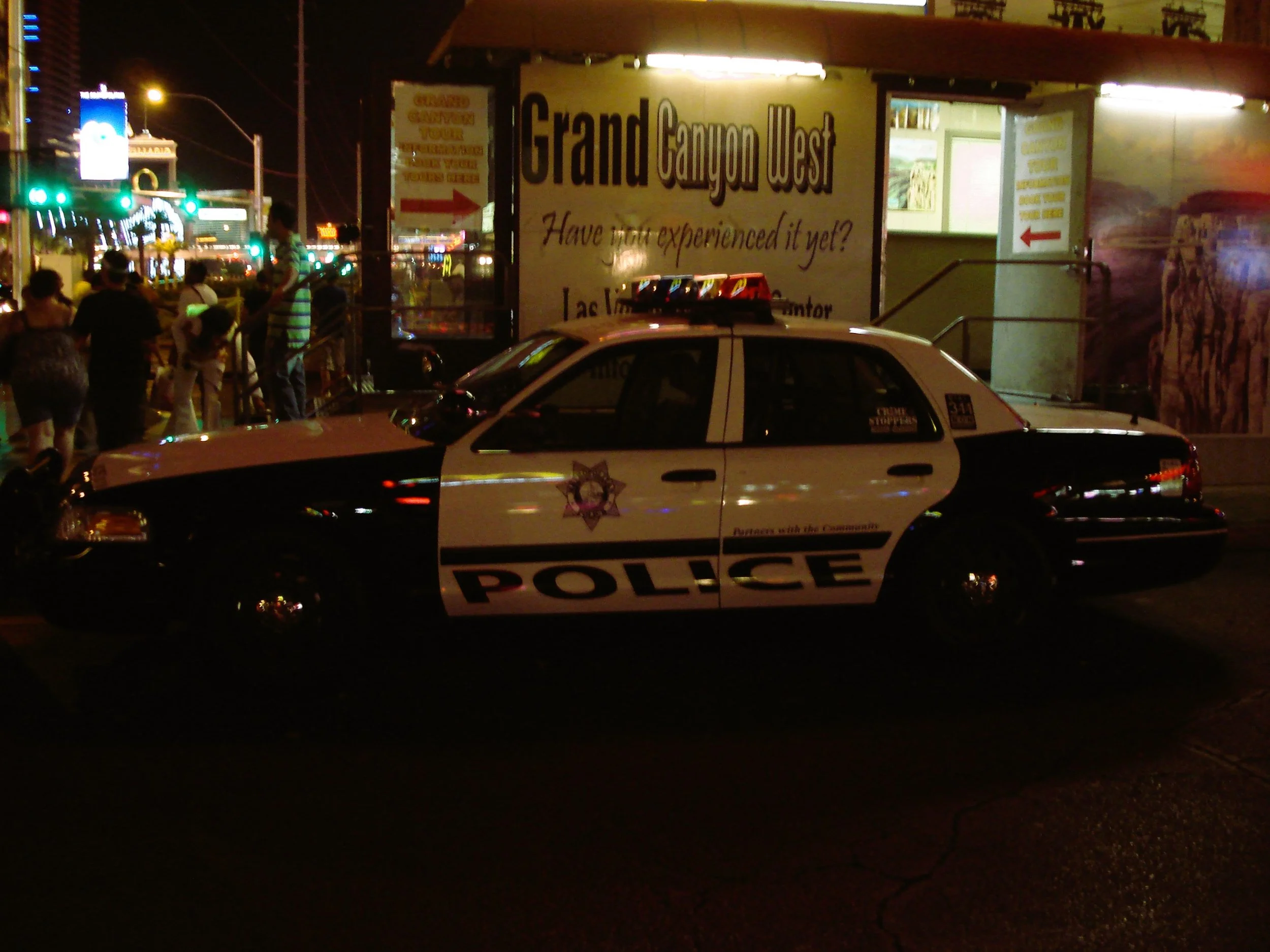 Un'auto della polizia parcheggiata di notte davanti a un'entrata con un cartellone che indica 'Grand Canyon West' e un gruppo di persone con luci e luci al neon sullo sfondo.