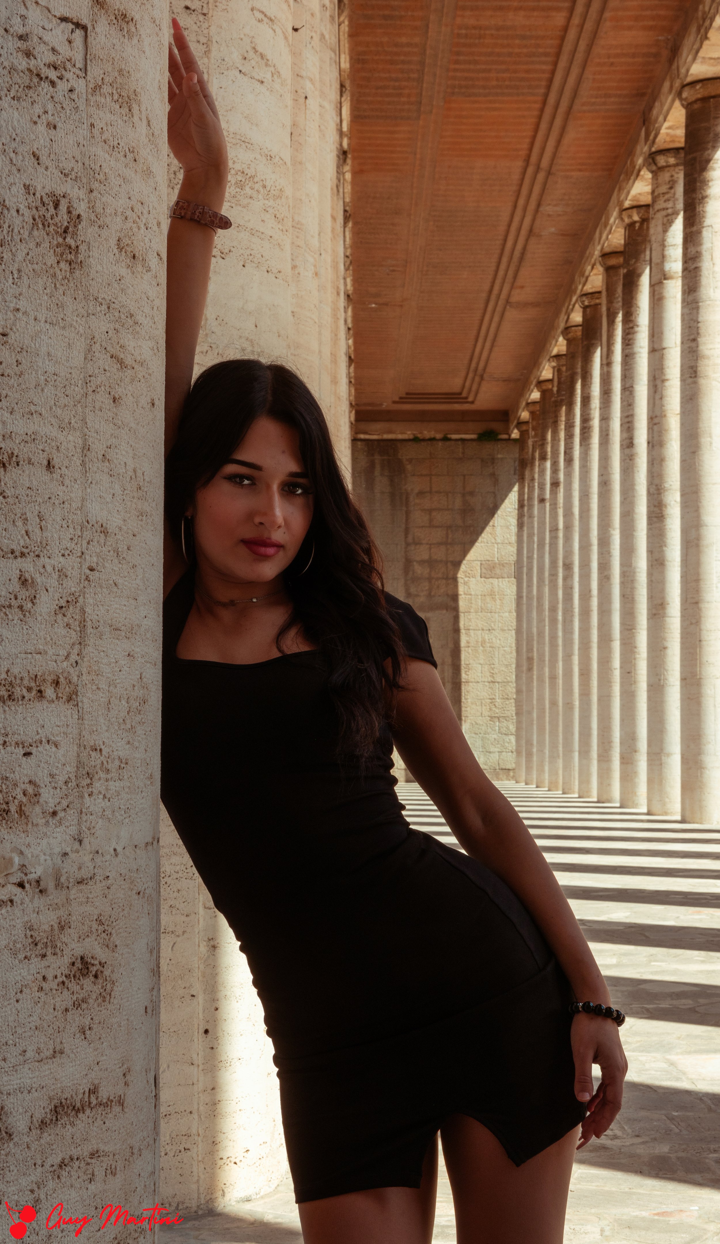 Una ragazza indossa un vestito nero e si appoggia contro una colonna in un'architettura antica con colonne in pietra e un soffitto in legno.