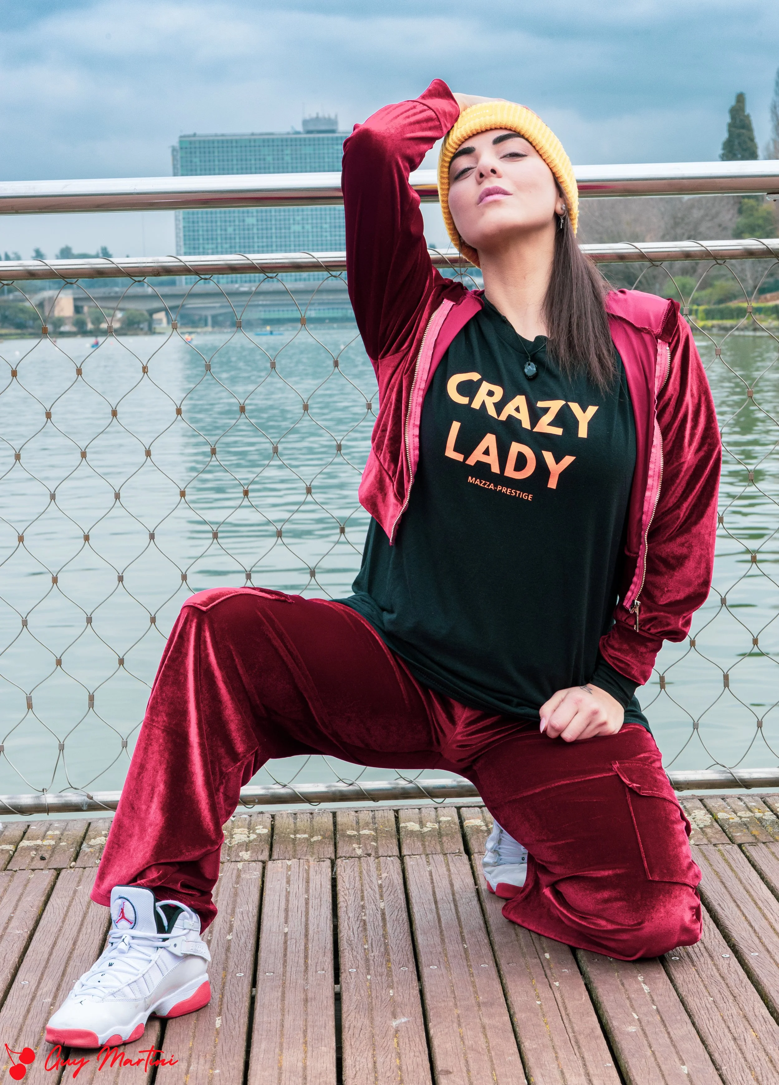 Una donna in abbigliamento sportivo con felpa e pantaloni di velluto rosso scuro, cappellino giallo, scarpe sportive bianche e neri si trova su un molo di legno accanto a un lago con un ponte barretta e un edificio moderno sullo sfondo, posa con uno 