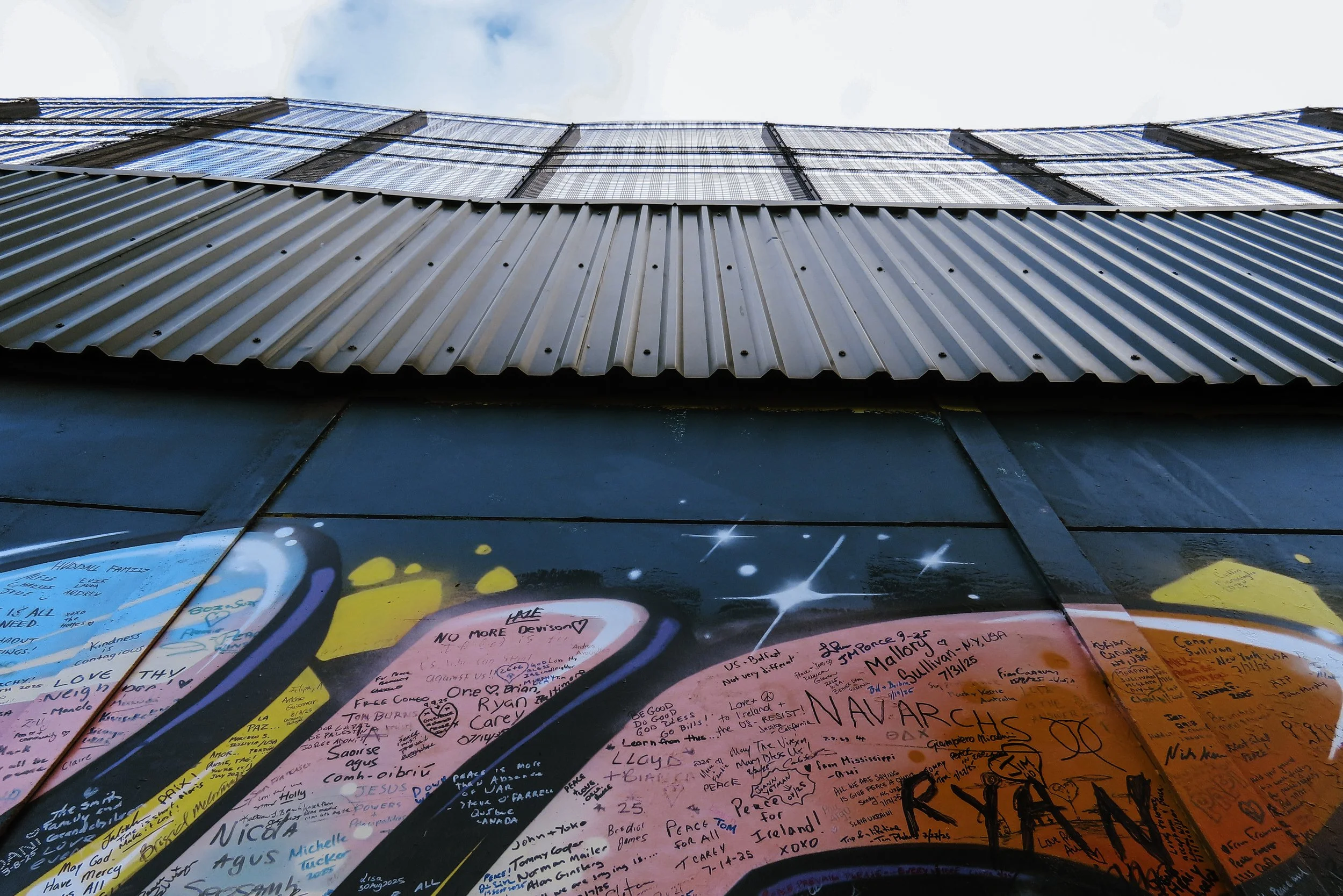 Murale colorata con scritte e messaggi di pace e amore, sotto un edificio con pannelli di vetro e tetto in metallo.