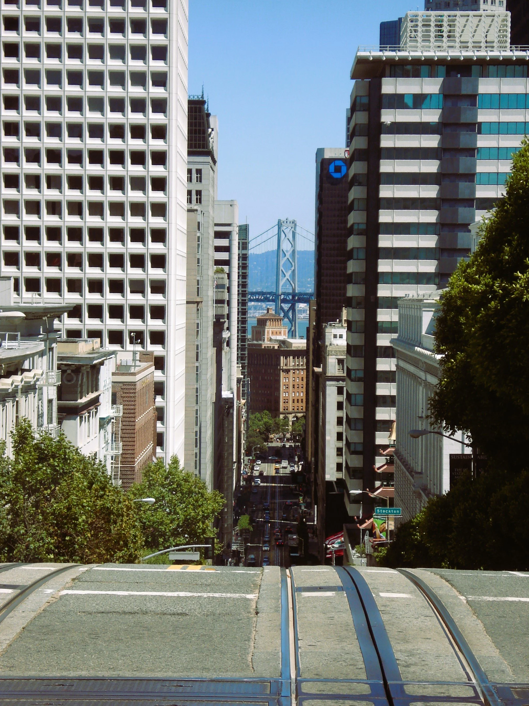 Vista di una strada di città con al centro un ponte sospeso in lontananza, circondata da edifici alti e moderne.

