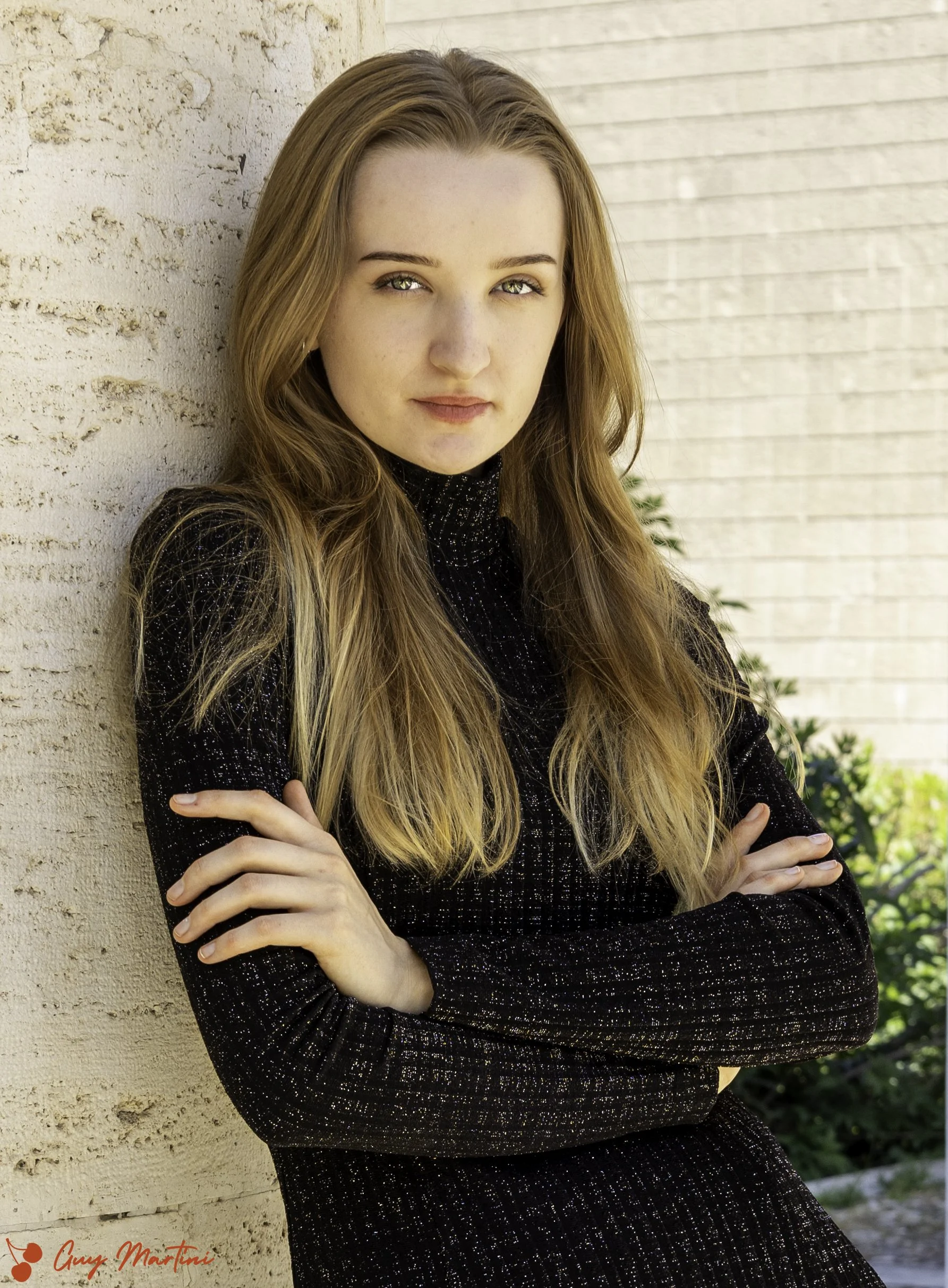 Ragazza con capelli lunghi e castani che si appoggia a un muro beige, indossando un abito nero a maniche lunghe, con un'espressione sicura e serena.