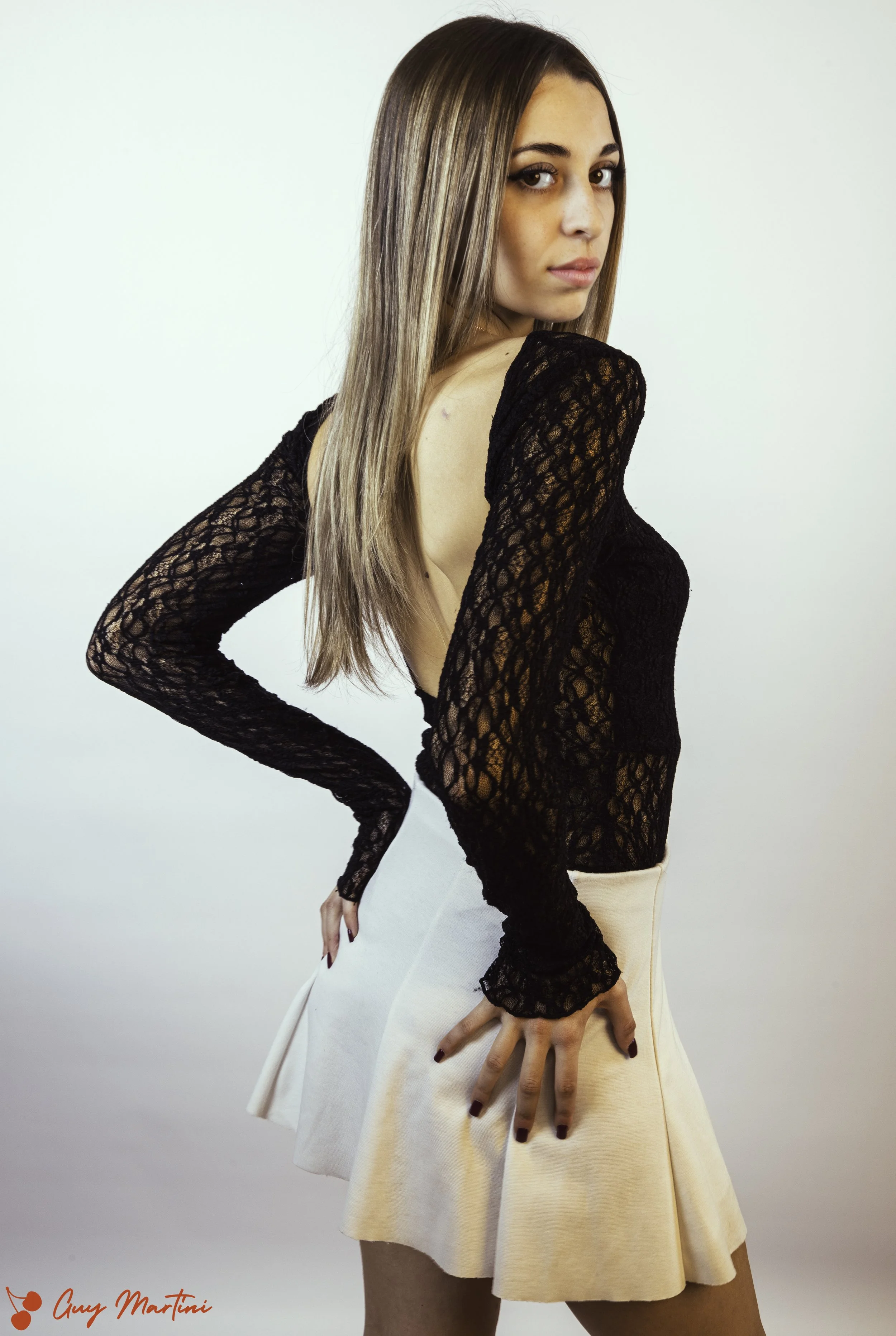 Ragazza con maglia nera in pizzo e gonna beige in piedi contro uno sfondo bianco, vista di profilo con sguardo rivolto verso la fotocamera.