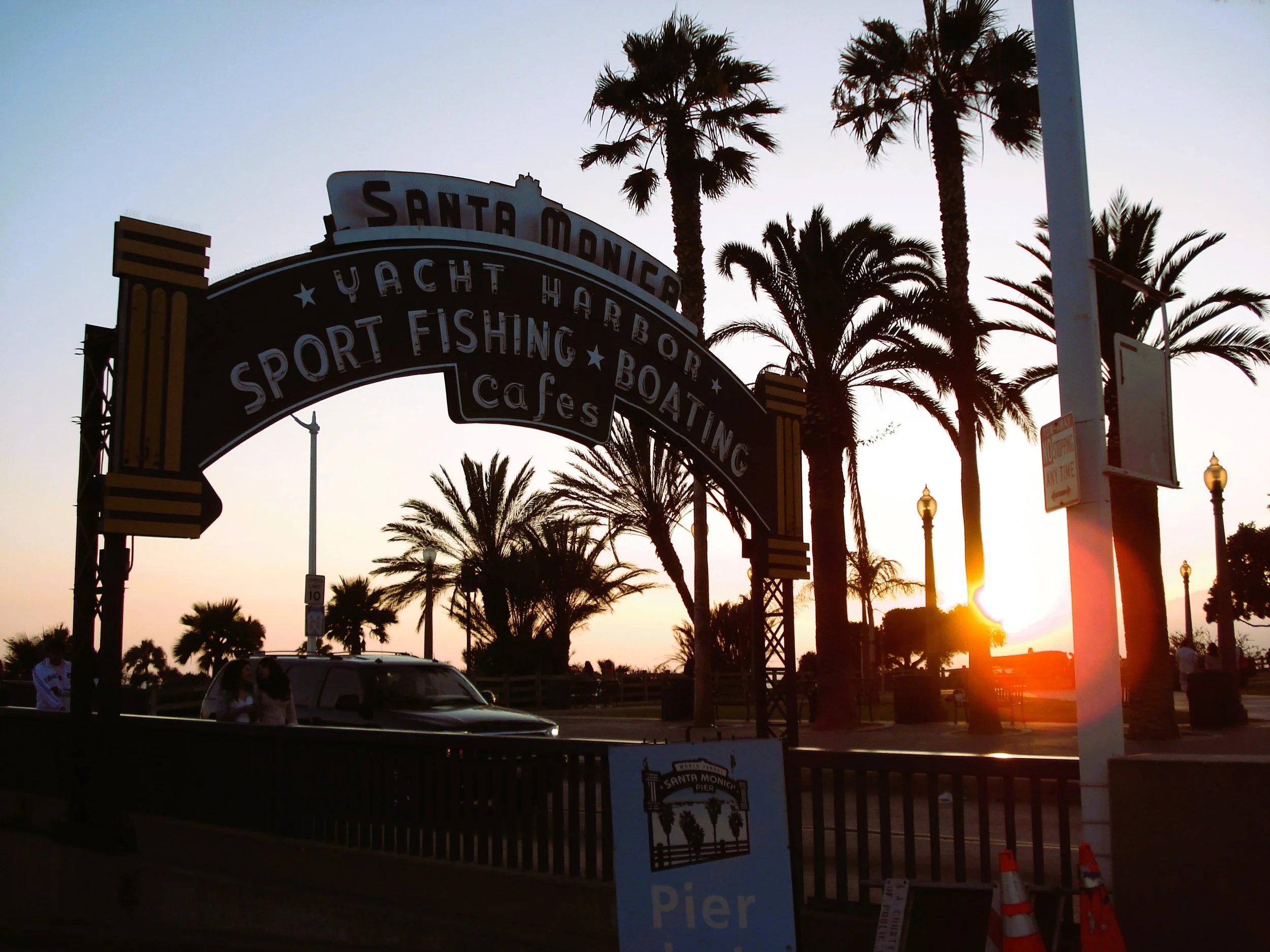Ingresso con cartello in legno e lettere luminose per Santa Monica Pier, con palme e lampioni al tramonto.