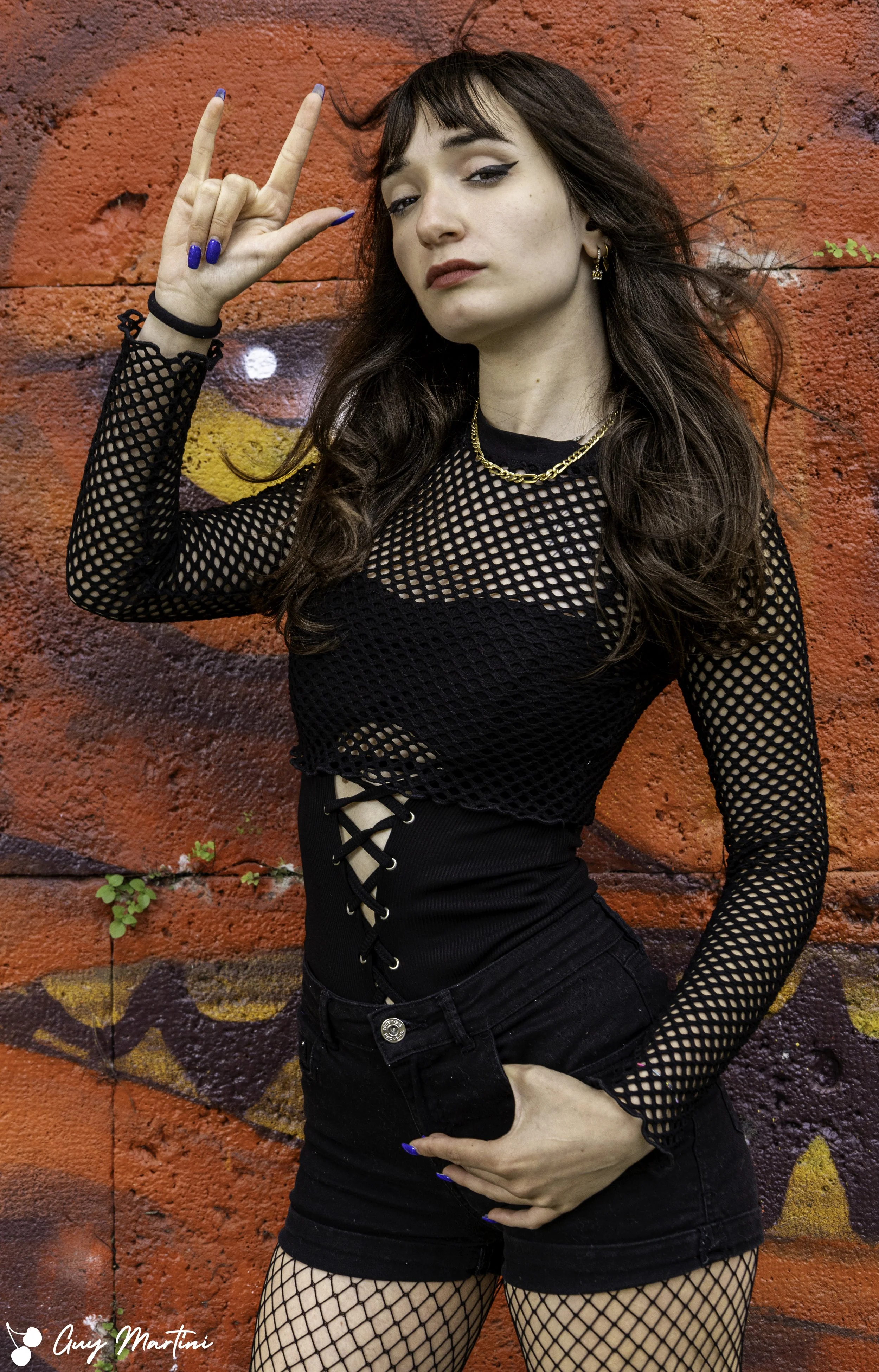 Una ragazza con capelli marroni, abbigliamento nero e accessori dorati, che fa il gesto della vittoria con le dita, davanti a un muro con graffiti rosso.