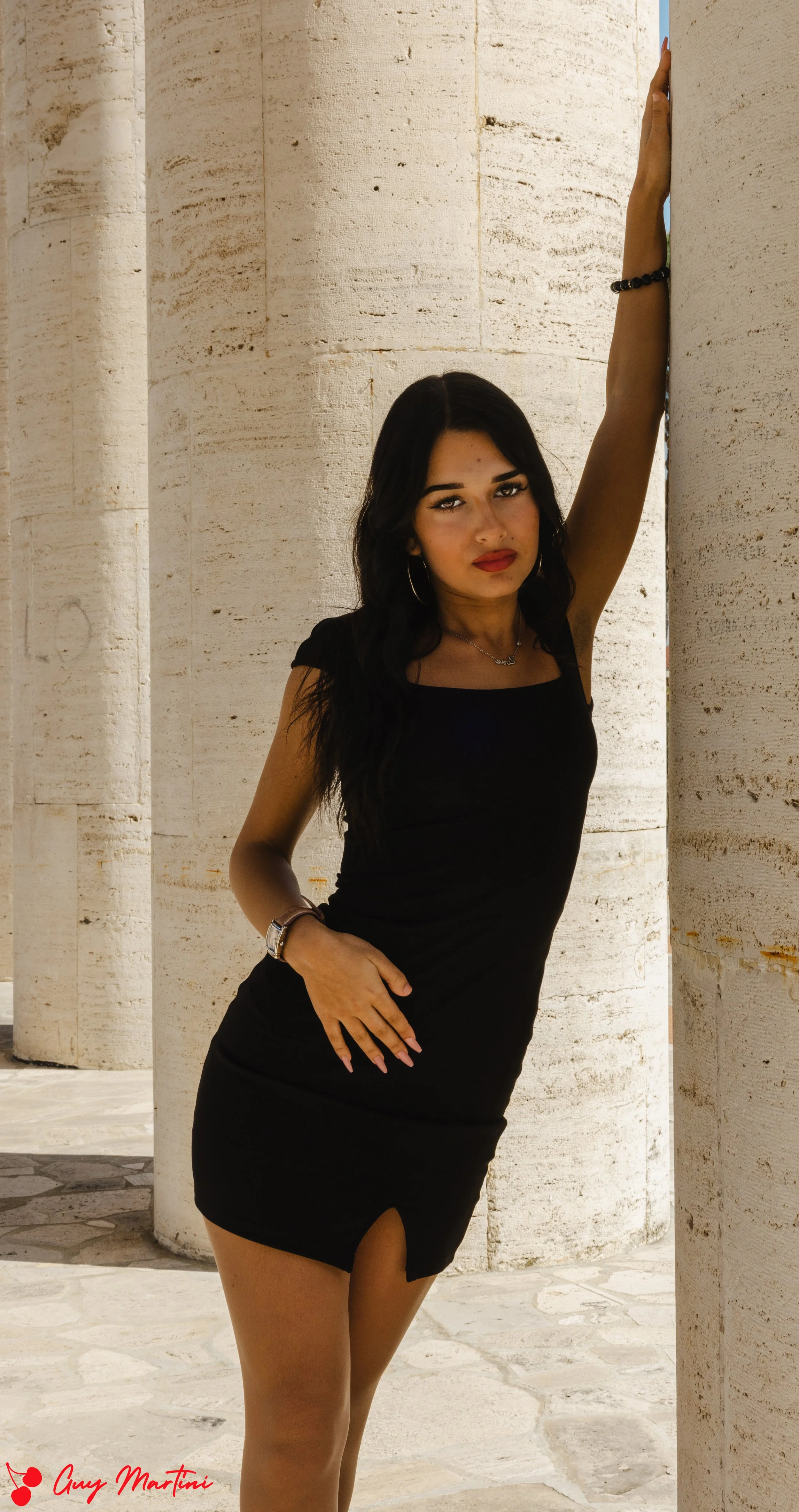 Una donna giovane con capelli neri e abbigliamento nero, che si appoggia a una colonna di pietra, con un ambiente esterno luminoso e colonne in pietra sullo sfondo.