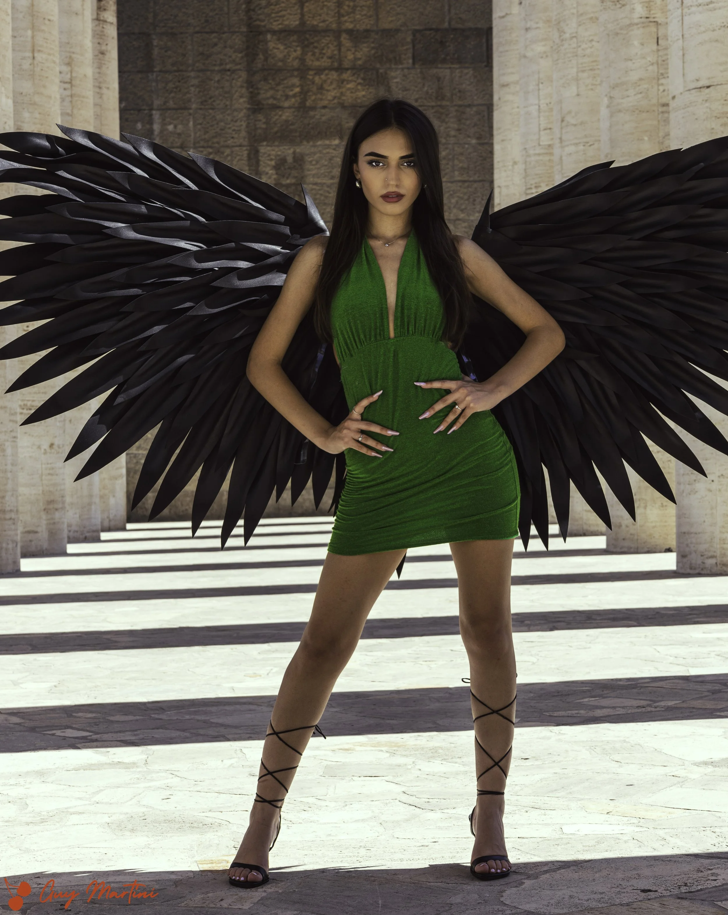 Una donna in abito verde brillante e scarpe nere con lacci incrociati, con grandi ali nere dietro di lei, si trova davanti a un edificio di pietra, in piedi con le mani sui fianchi.