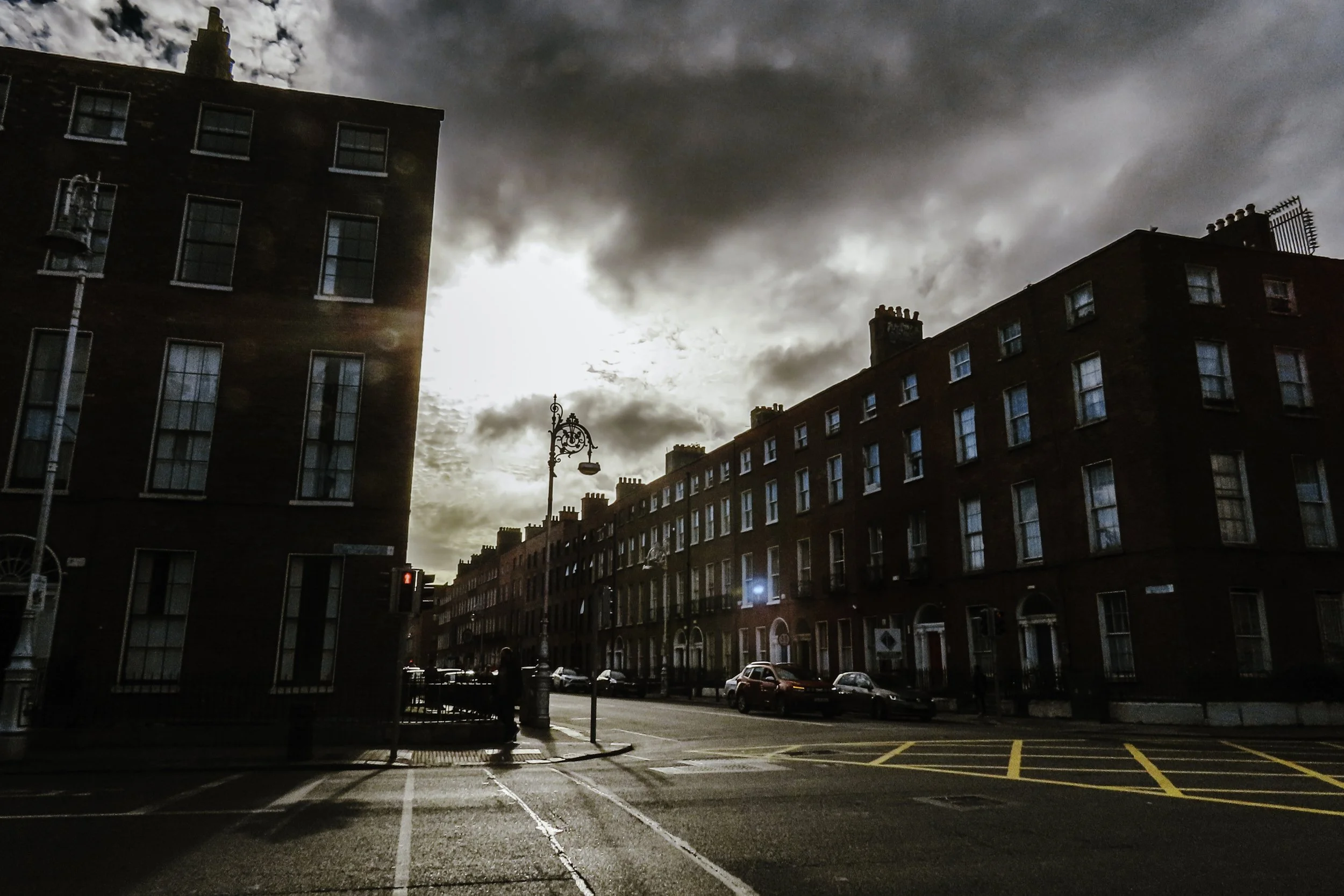 Immagine di una strada urbana con edifici in mattoni, auto parcheggiate e lampioni, sotto un cielo nuvoloso con il sole che tramonta.