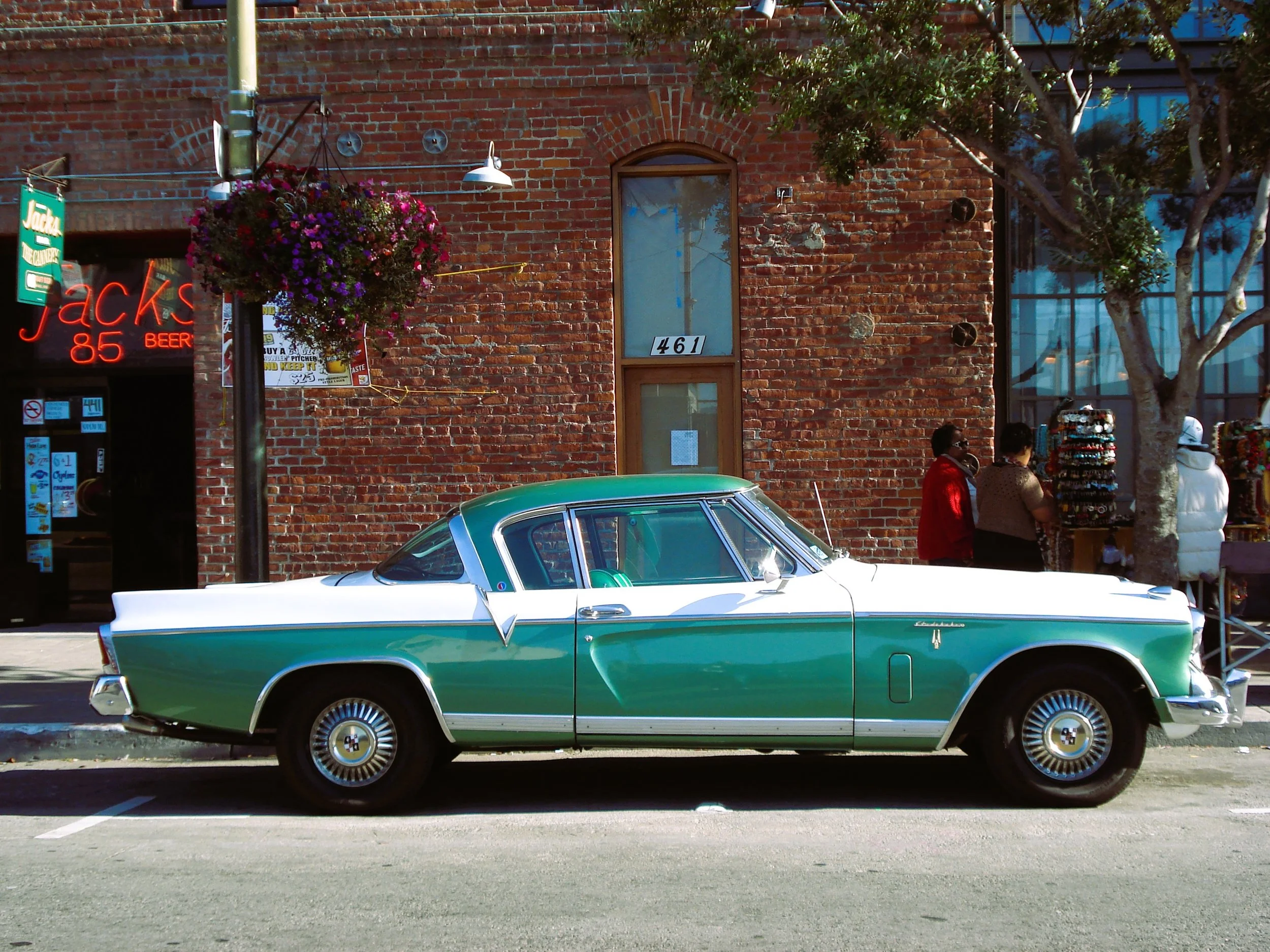 Automobile vintage verde e bianca parcheggiata lungo il marciapiede davanti a un edificio in mattoni rossi, con persone che passeggiano e negozi visibili sullo sfondo.
