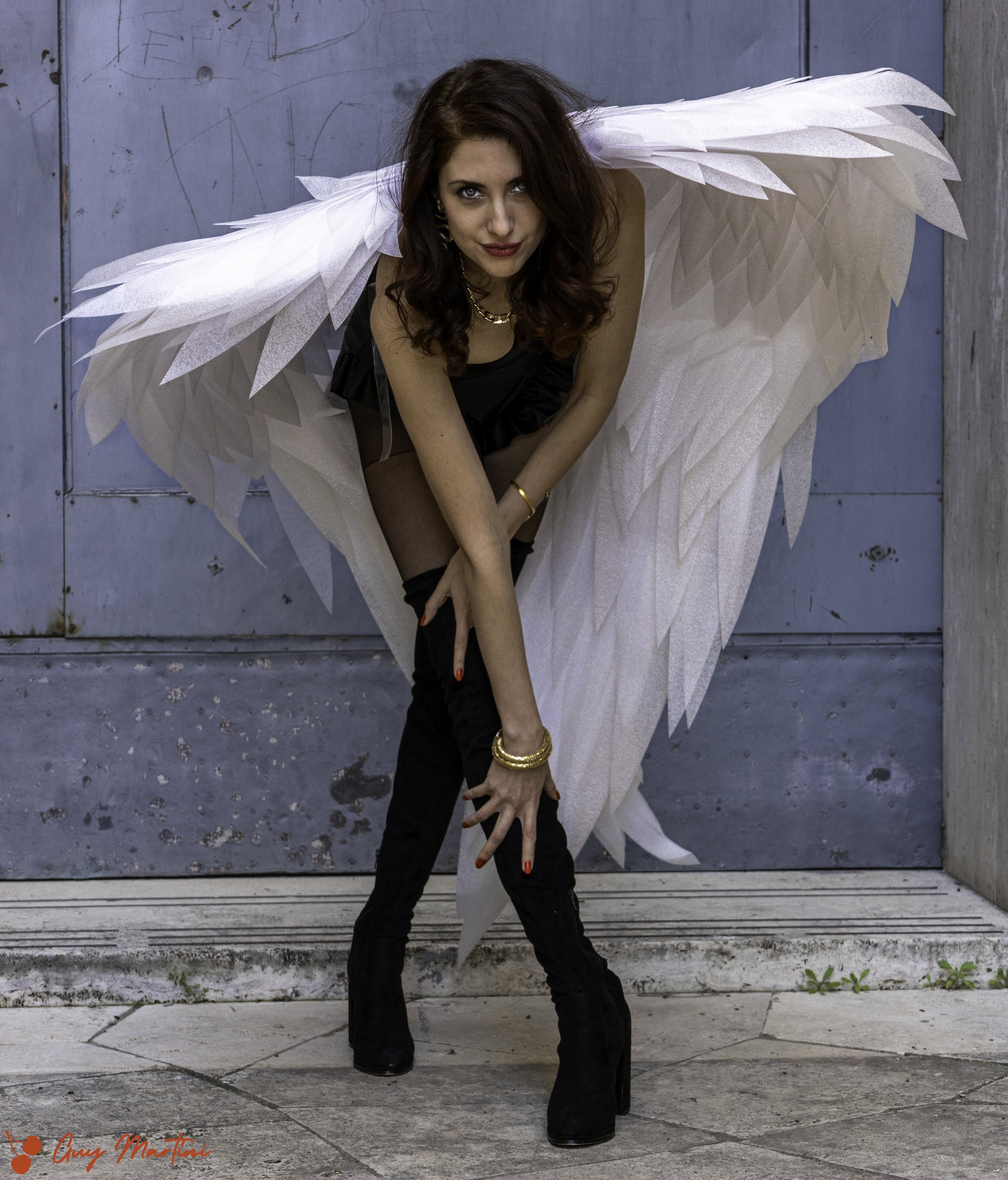 Una donna con ali biene, indossa abbigliamento nero e stivali alti neri, posa in punta di piedi.