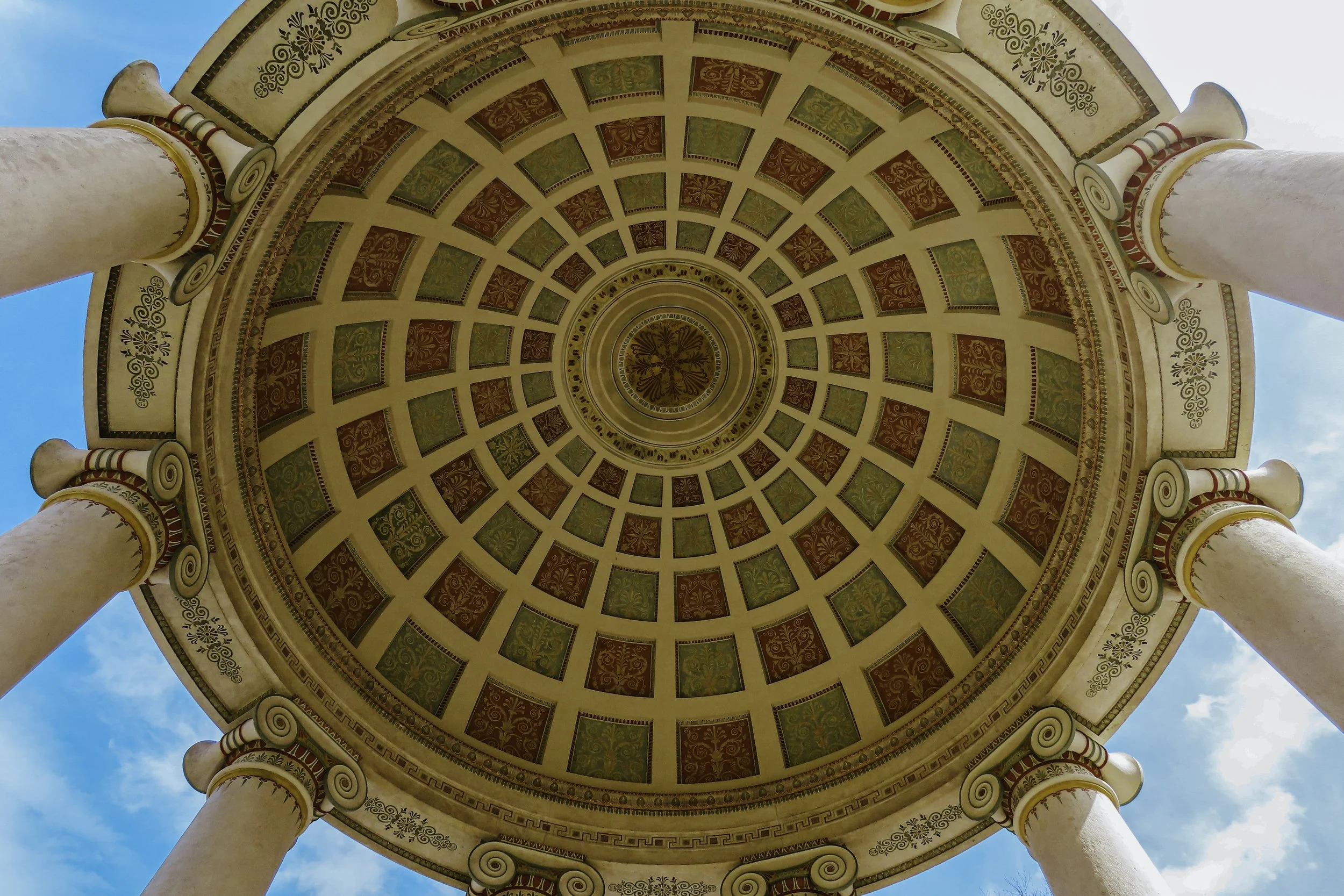 Vista dal basso di un soffitto a cupola decorato con motivi geometrici e colonne corinzie, con il cielo azzurro sullo sfondo.