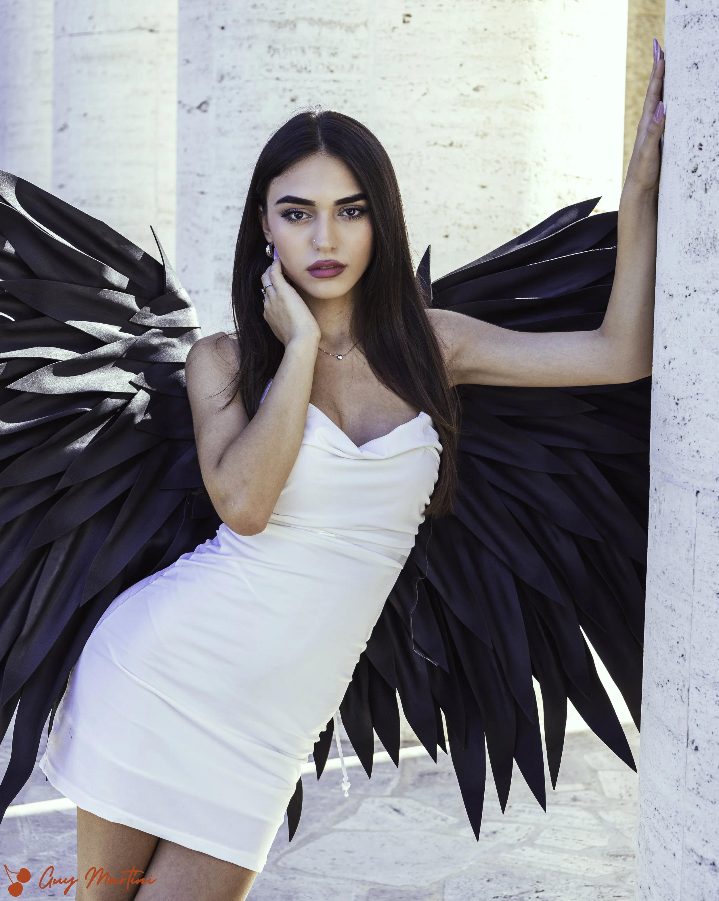 Una donna giovane con lunghi capelli castani, indossa un vestito bianco aderente e ha grandi ali nere da farfalla. Sembra essere in una posa artistica vicino a un muro di cemento.