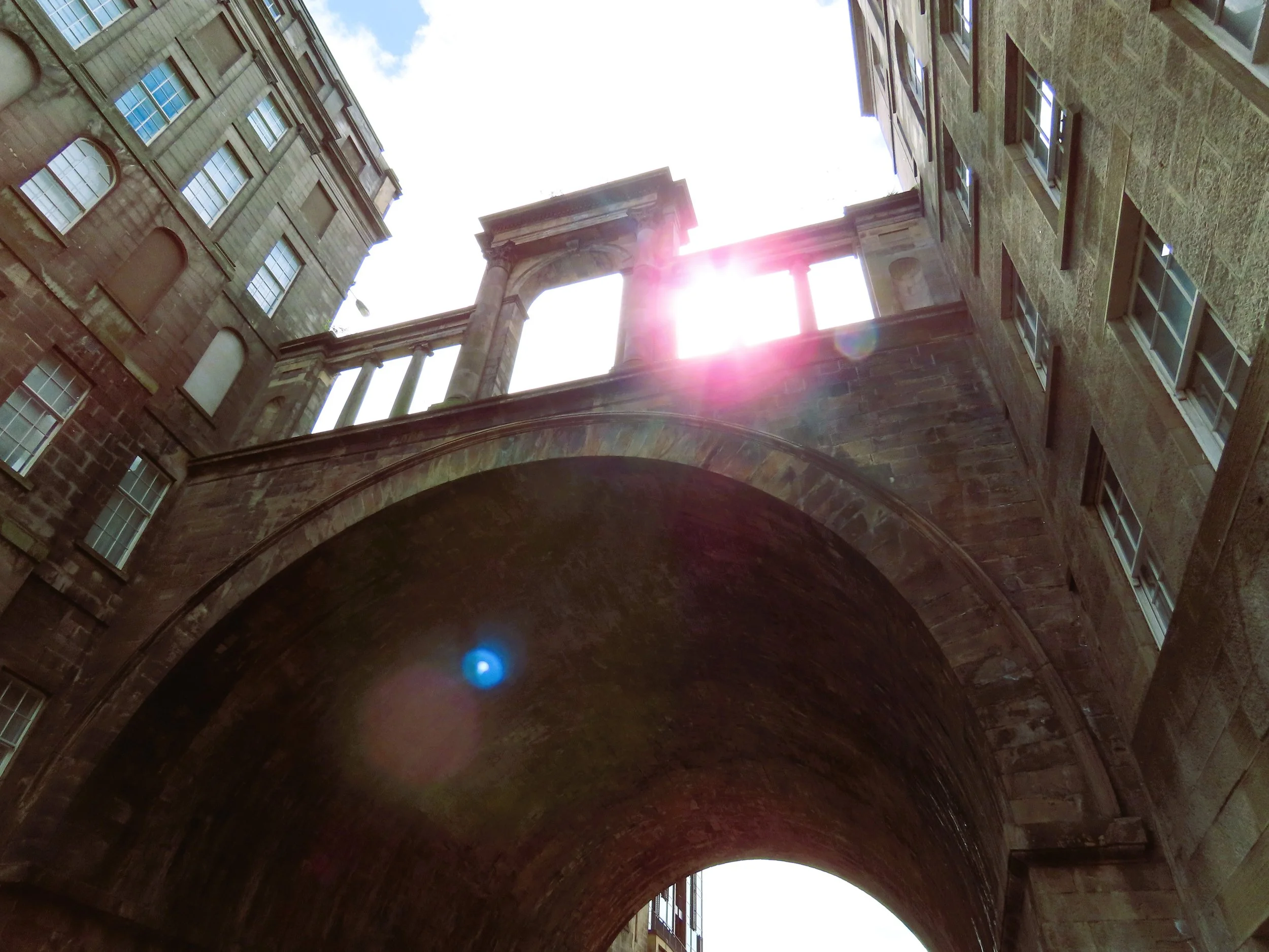 Immagine di un arco in pietra che collega due edifici storici con finestre, sotto uno scorcio del cielo con il sole.