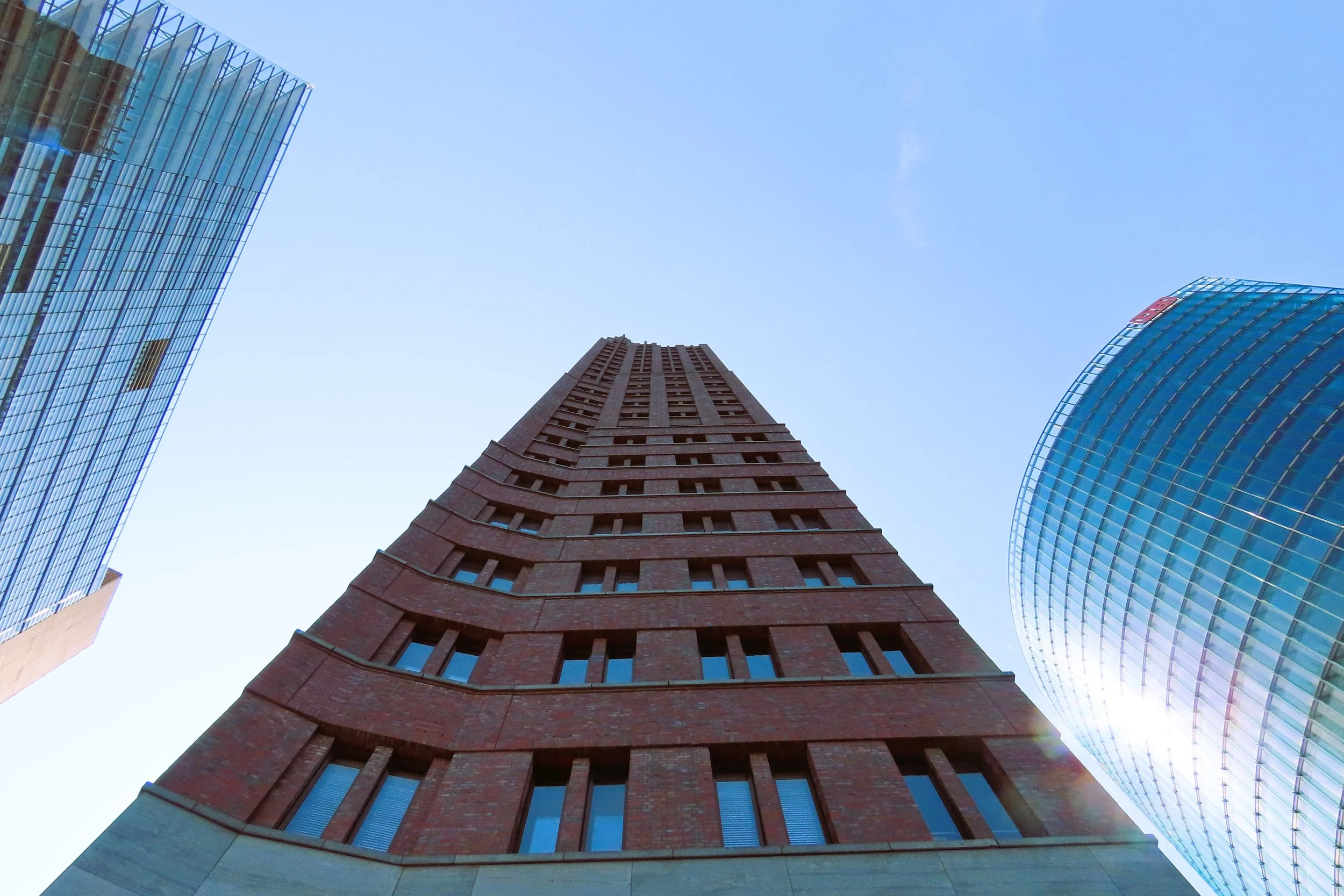 Immagine aerea di un grattacielo in mattoni rossi con finestre piccole e ordinate, circondato da altri edifici moderni in vetro, sotto un cielo azzurro senza nuvole.