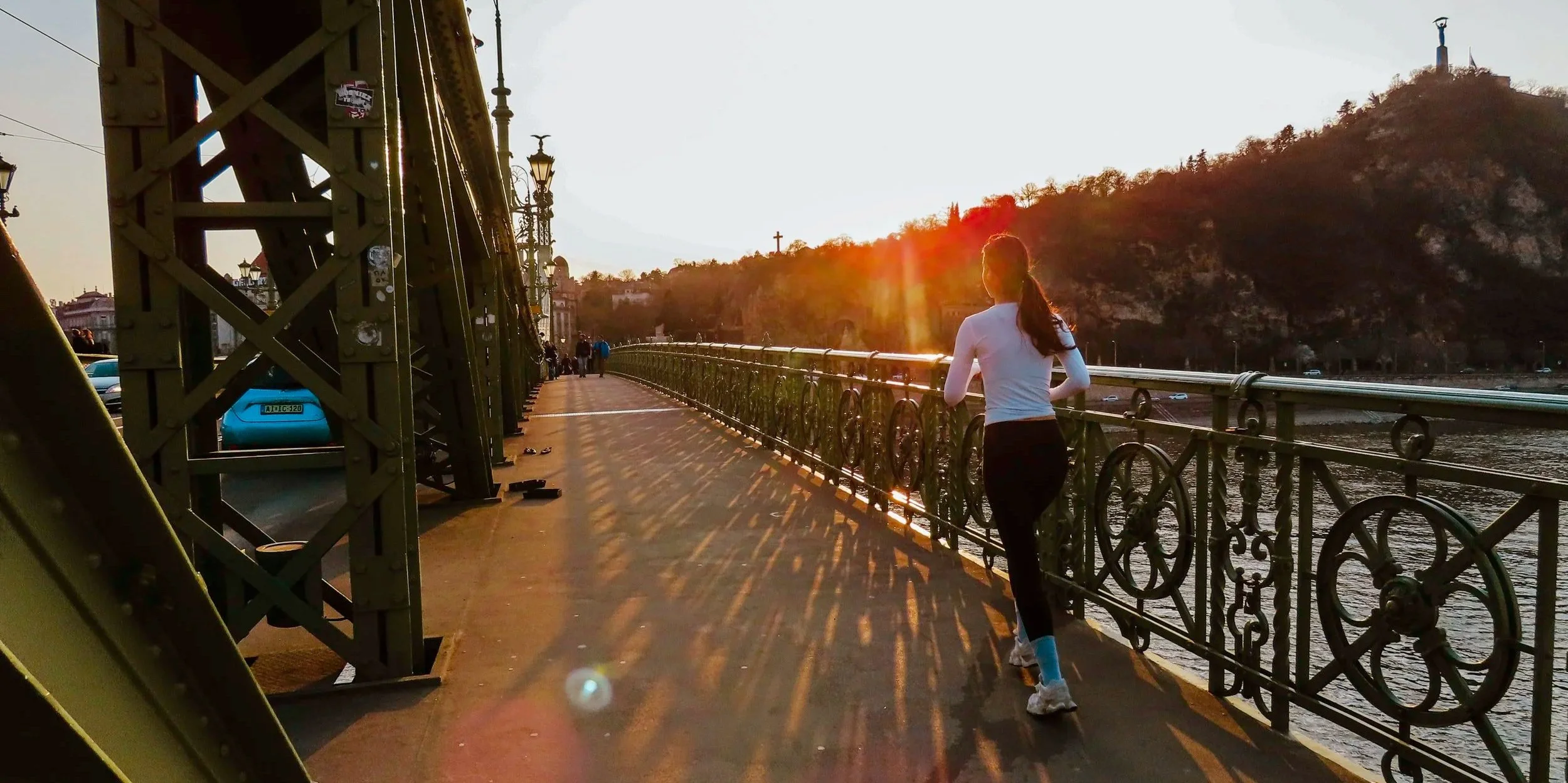 Una donna corre lungo un ponte con ringhiere di ferro, al tramonto, con una collina e una croce sullo sfondo. Il ponte ha lampioni e ci sono alcune persone in lontananza.