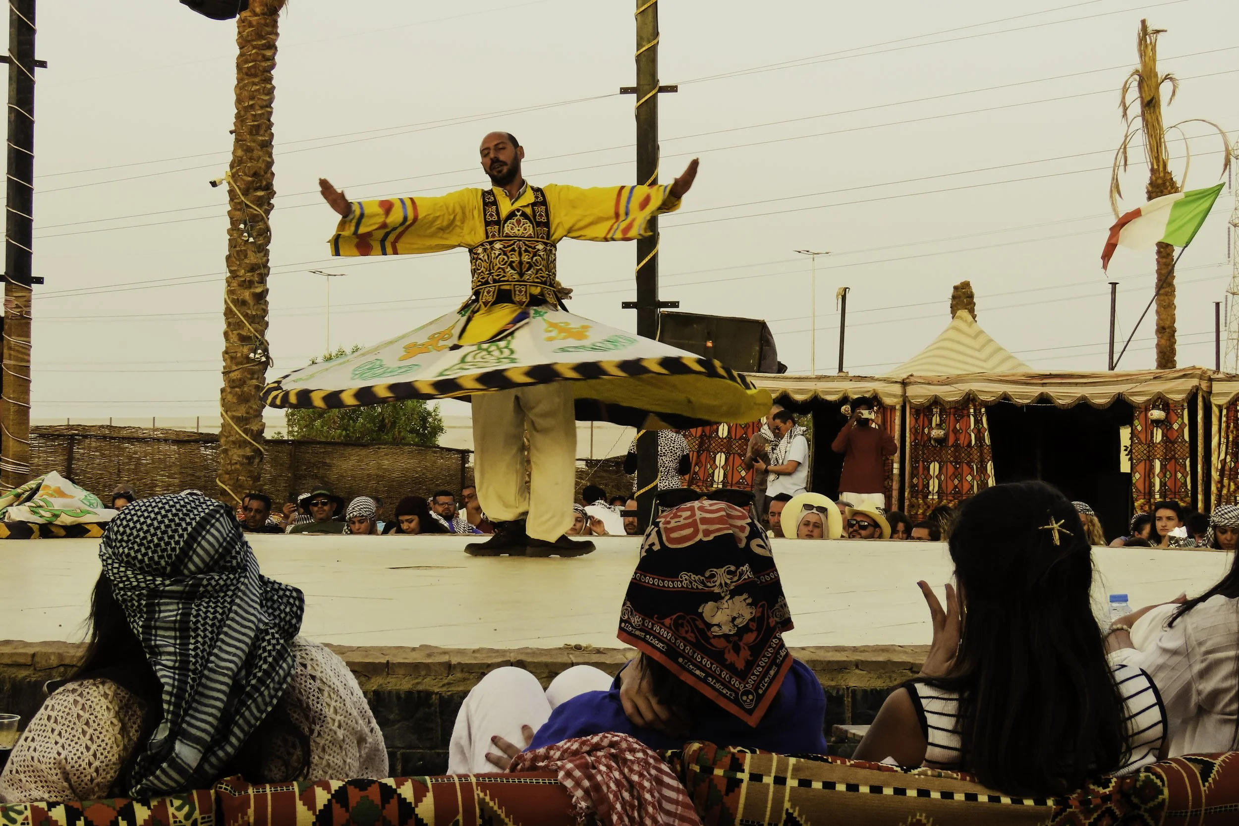 Artista che danza durante uno spettacolo tradizionale con pubblico seduto di fronte, vestito con abiti colorati e accessori tradizionali, sotto tende decorative e palme.