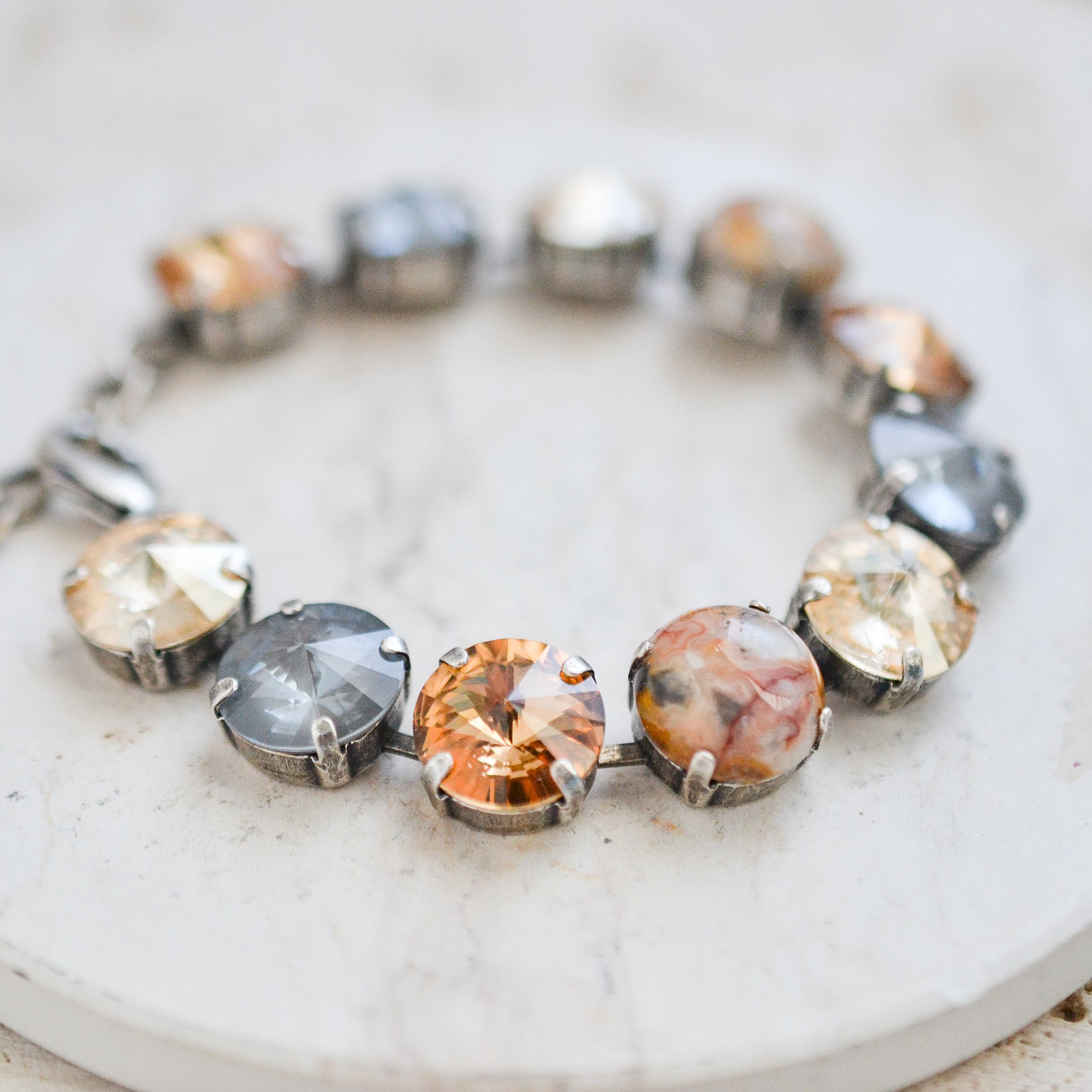 Close-up of a silver bracelet with large, round, faceted gemstone beads in various shades of gray, orange, and marbled patterns, resting on a white stone surface.